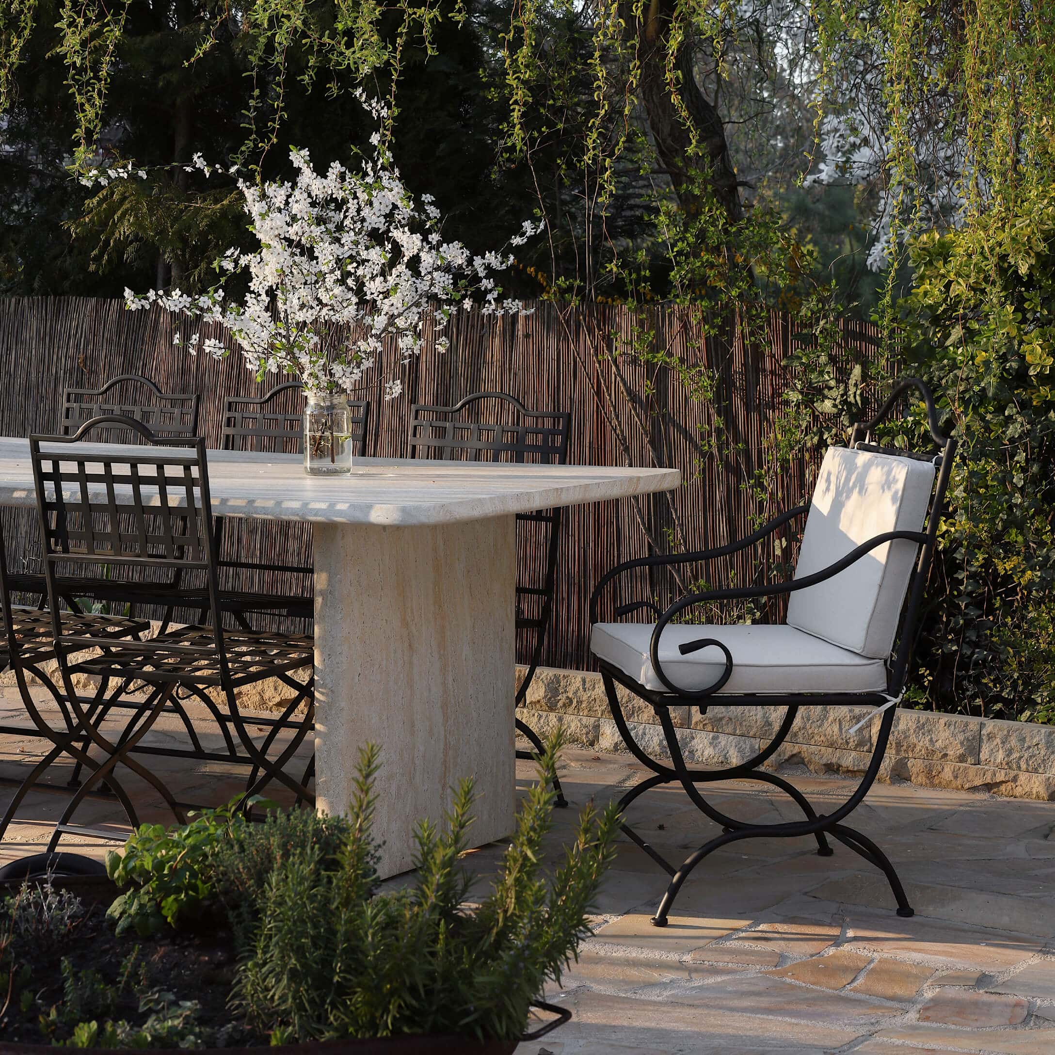 Table en pierre avec chaises en métal noir et coussins clairs dans un jardin fleuri