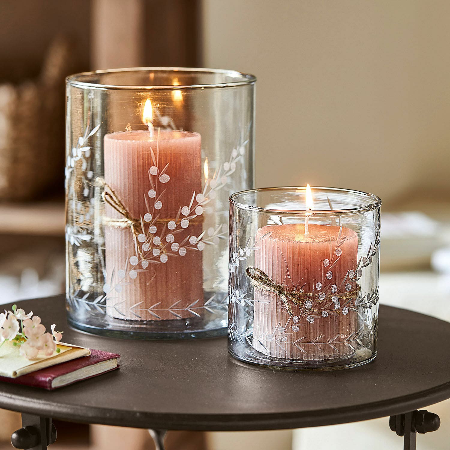 Lot de 2 photophores Temao Deux bougies roses dans des photophores en verre transparent avec motif végétal blanc sur une table