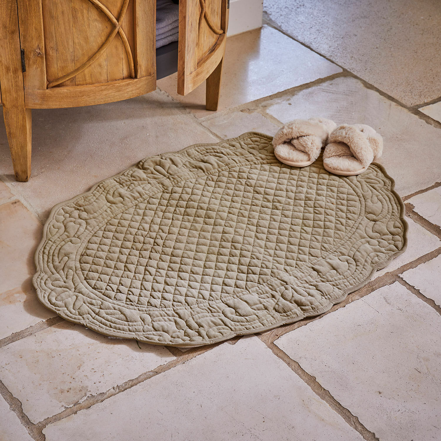 Tapis de bain Courlevin Tapis matelassé ovale en coton couleur olive sur sol en pierre avec chaussons beiges à côté