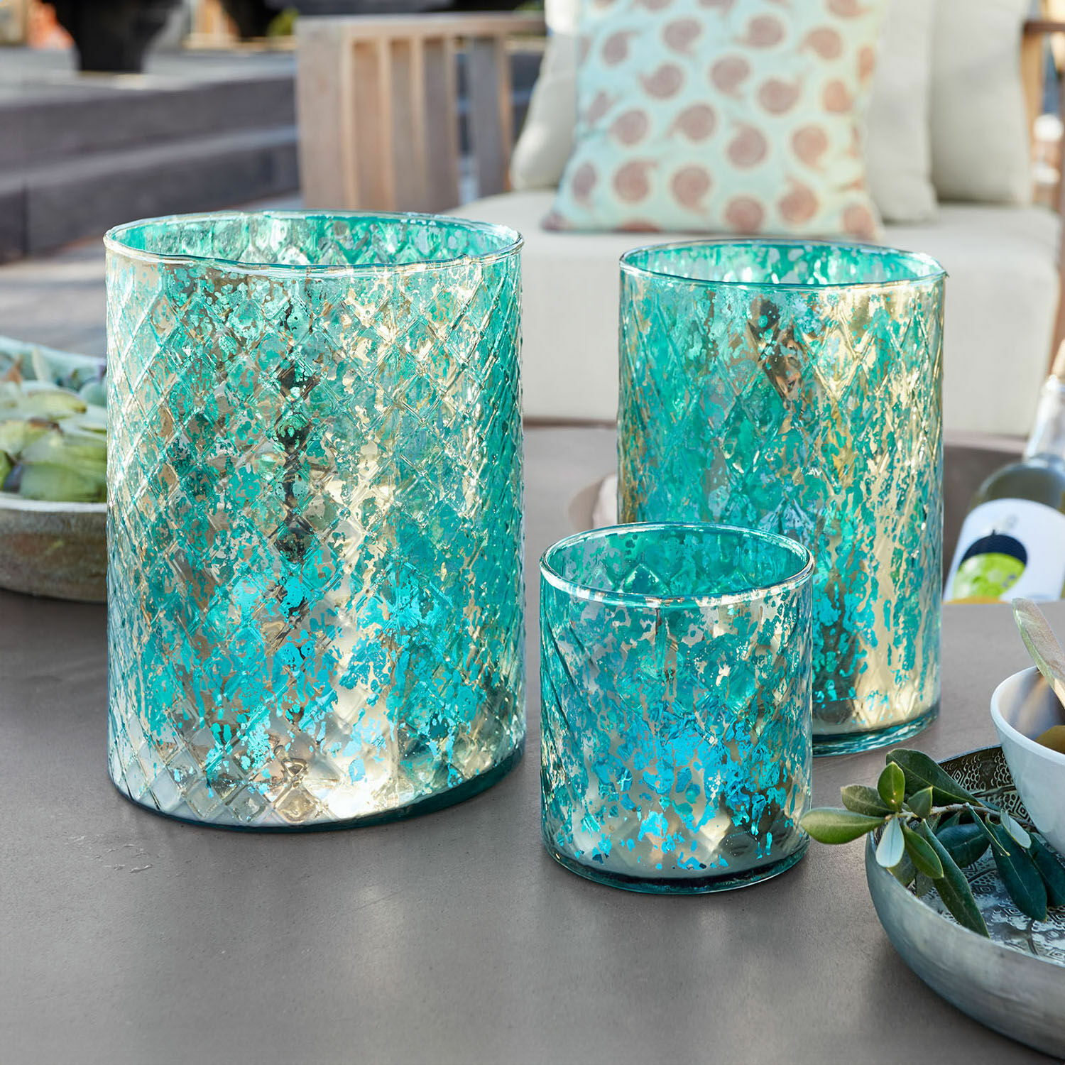 Lot de 3 photophores Firalen Ensemble de trois photophores en verre turquoise avec motif métallique sur une table extérieure
