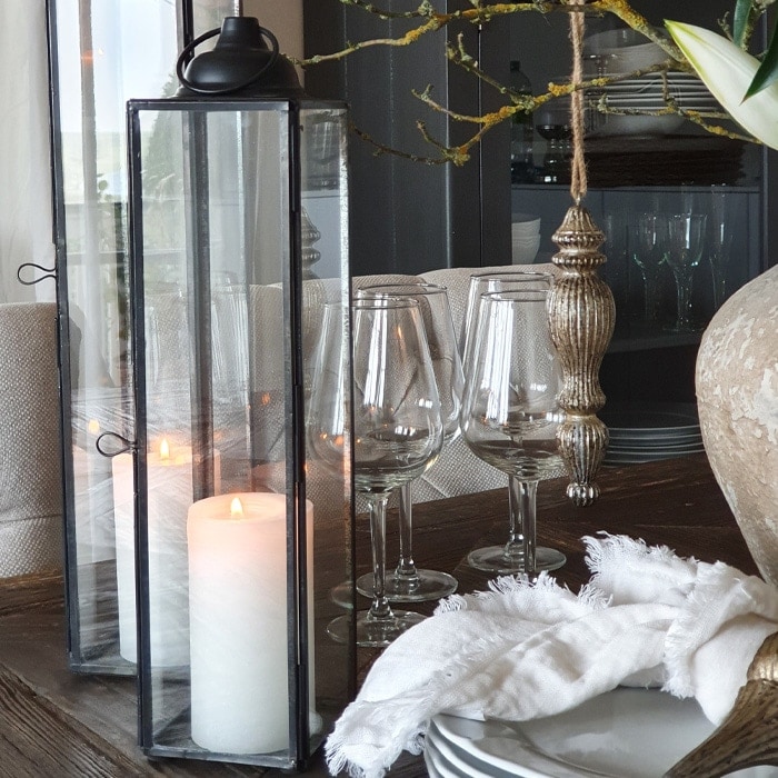 Lanterne en verre avec cadre métallique noir et bougie blanche sur table en bois avec verres et tissu blanc