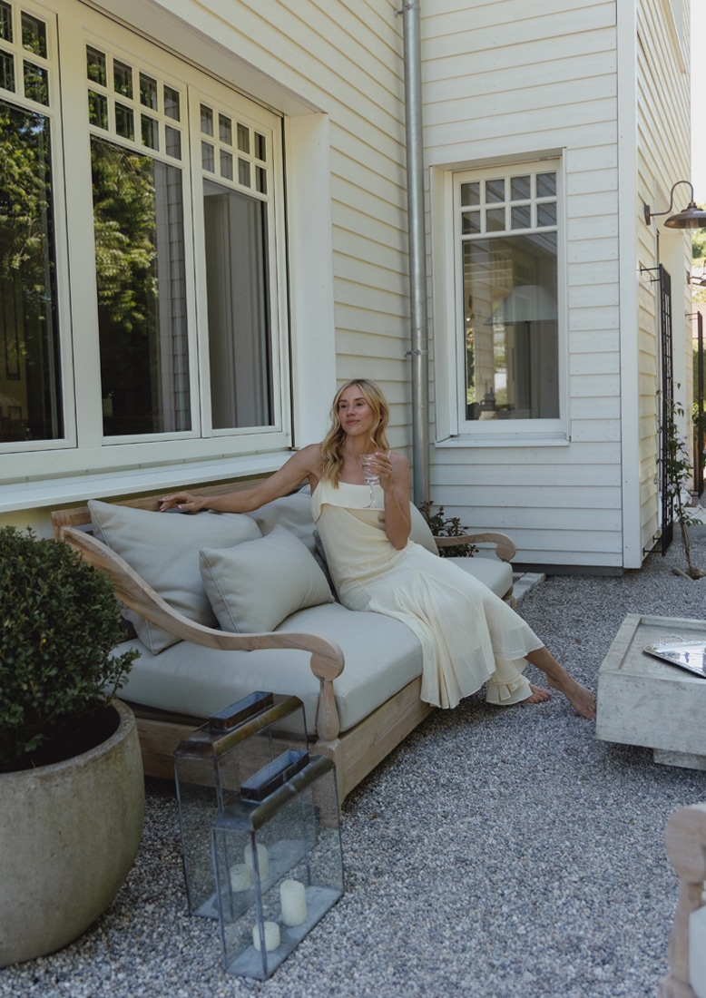Canapé de jardin en bois avec coussins gris clair sur patio en gravier avec femme en robe crème