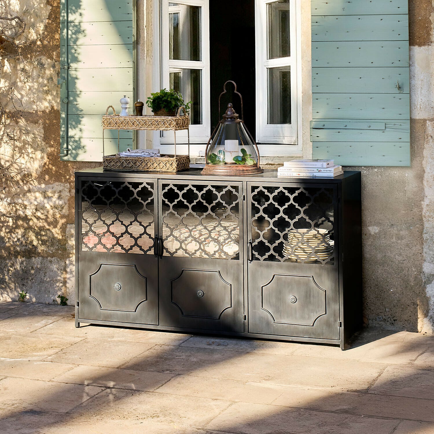 Buffet bas Astralon Buffet noir en métal avec portes ajourées sur une terrasse devant une fenêtre aux volets verts