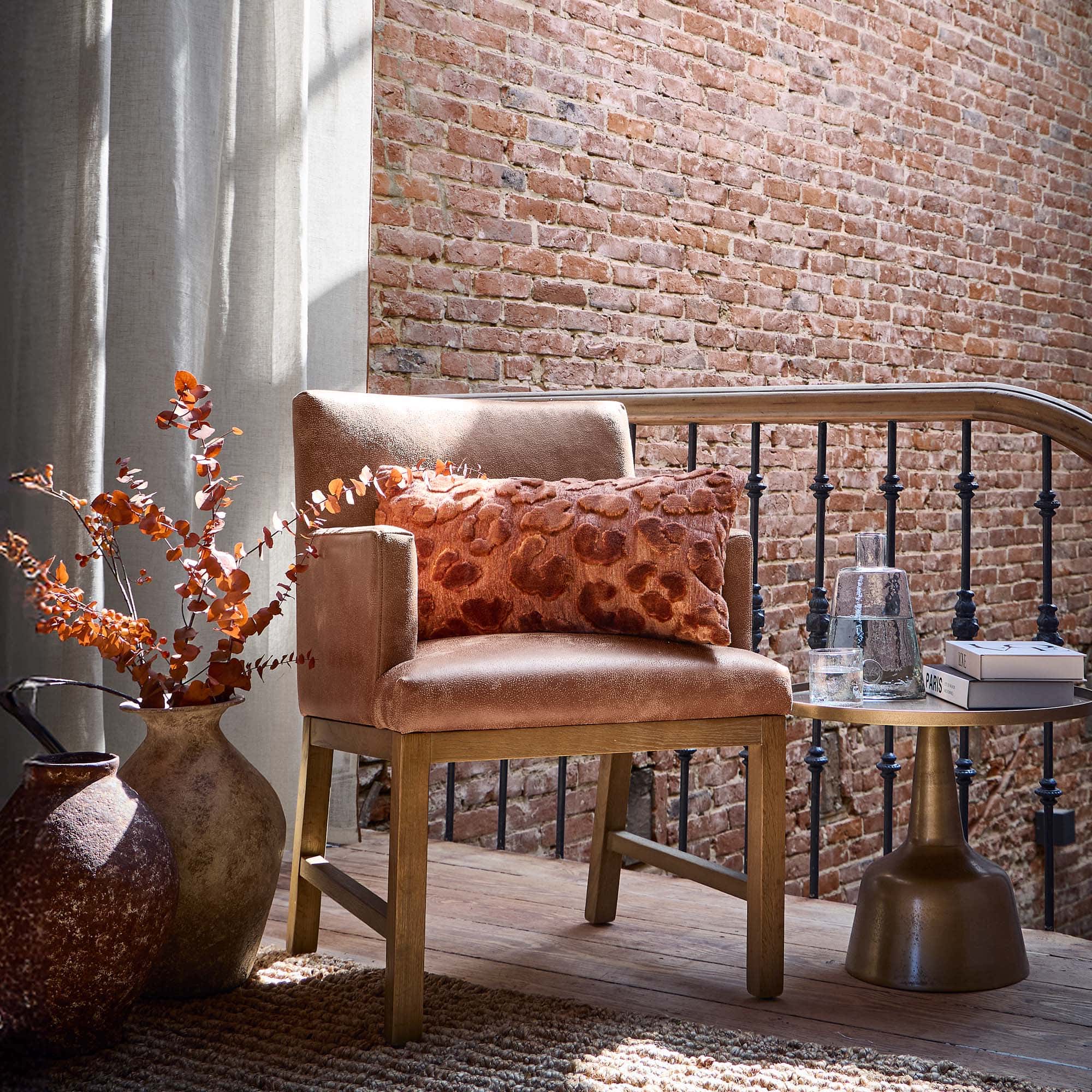 Fauteuil beige avec base en bois et coussin orange devant un mur en briques et des rideaux