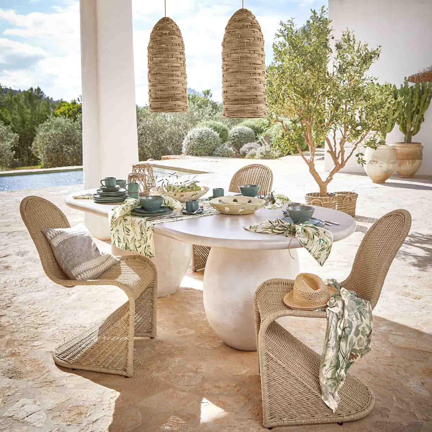 Table ovale blanche en pierre avec chaises en osier et suspensions naturelles dans un jardin ensoleillé