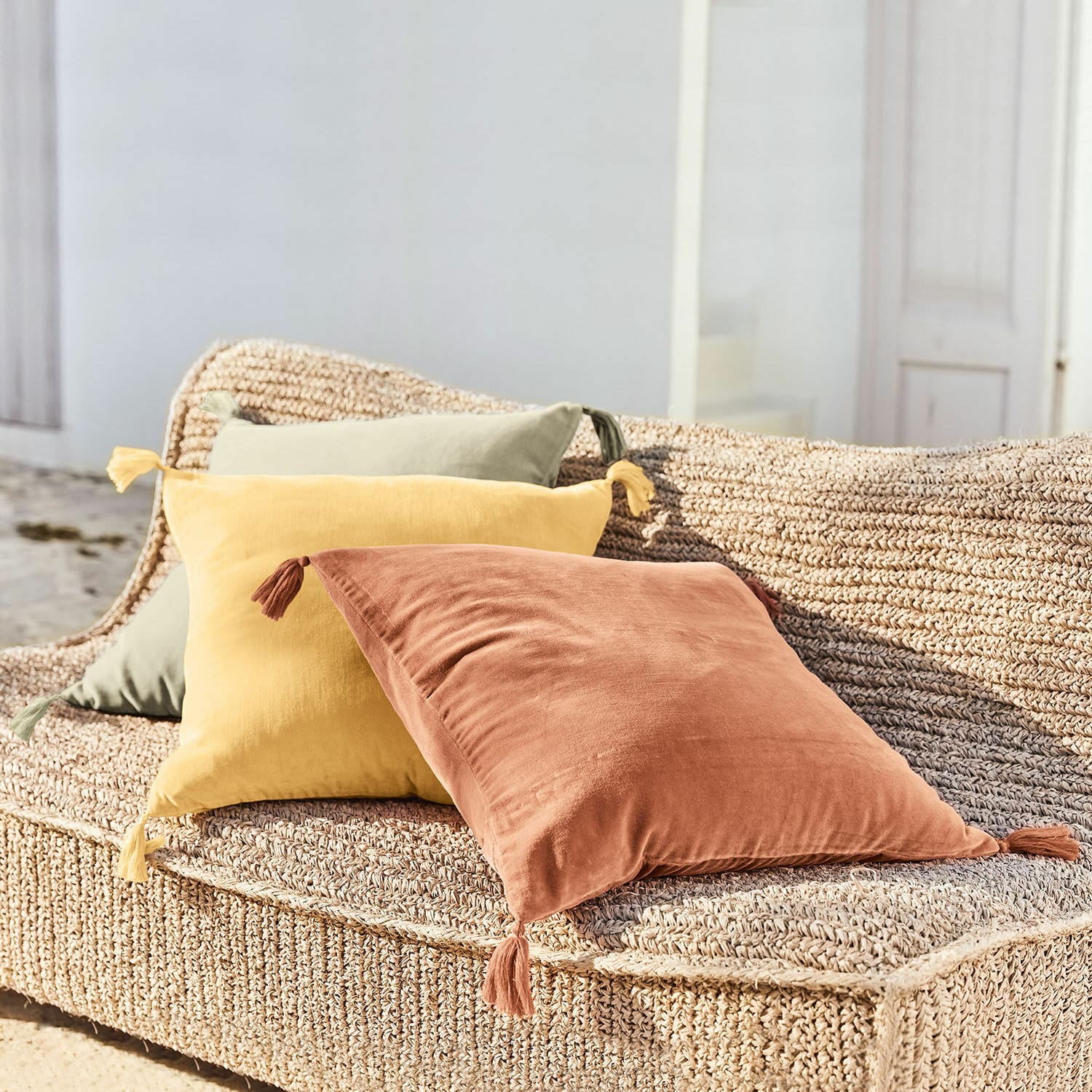 Trois coussins décoratifs en coton terracotta, jaune et vert sur un siège tressé en extérieur