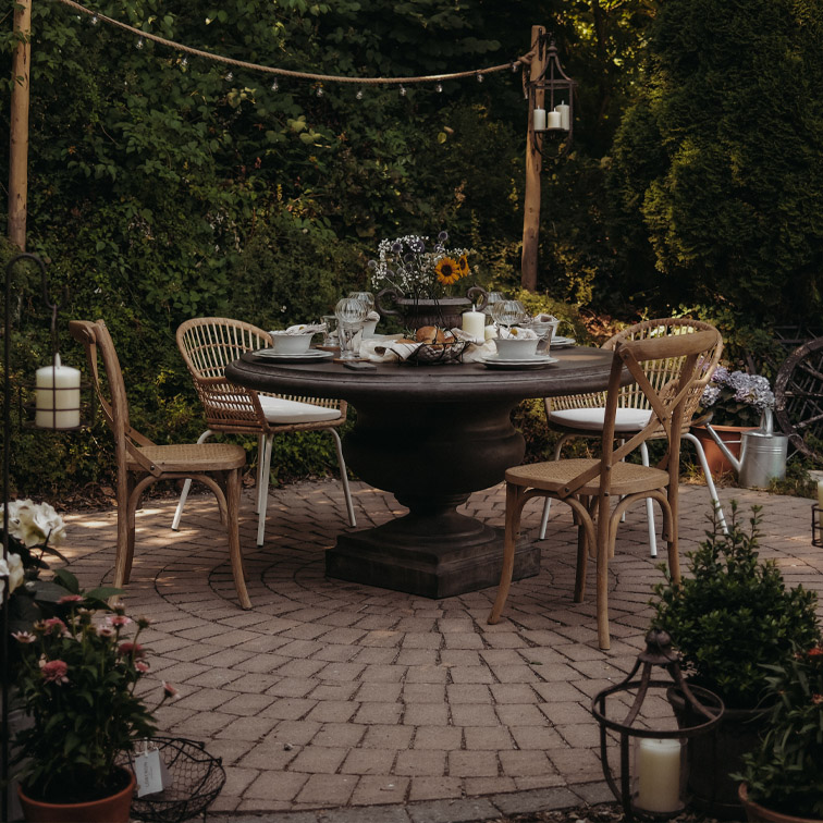 Table ronde en bois foncé avec chaises en osier sur terrasse pavée décorée de bougies et fleurs en extérieur