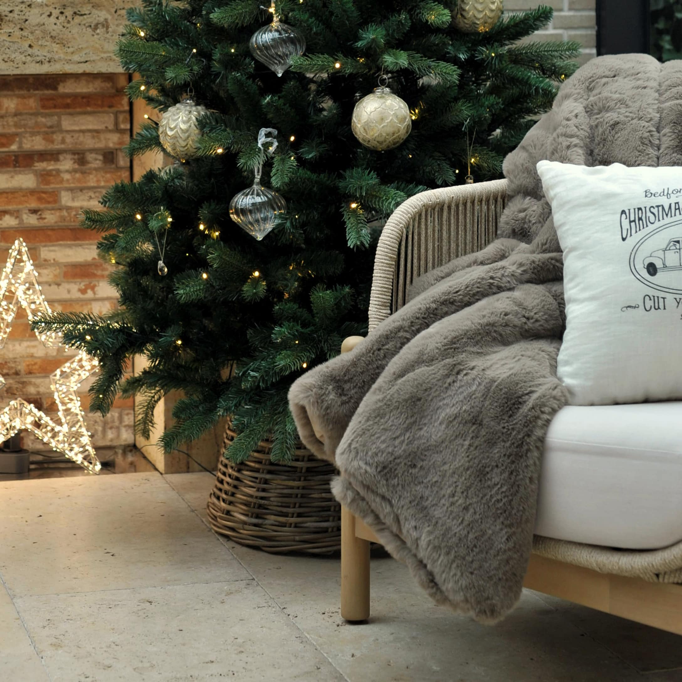 Coin cosy avec plaid en fausse fourrure grise et coussin blanc près d'un sapin décoré et étoile lumineuse
