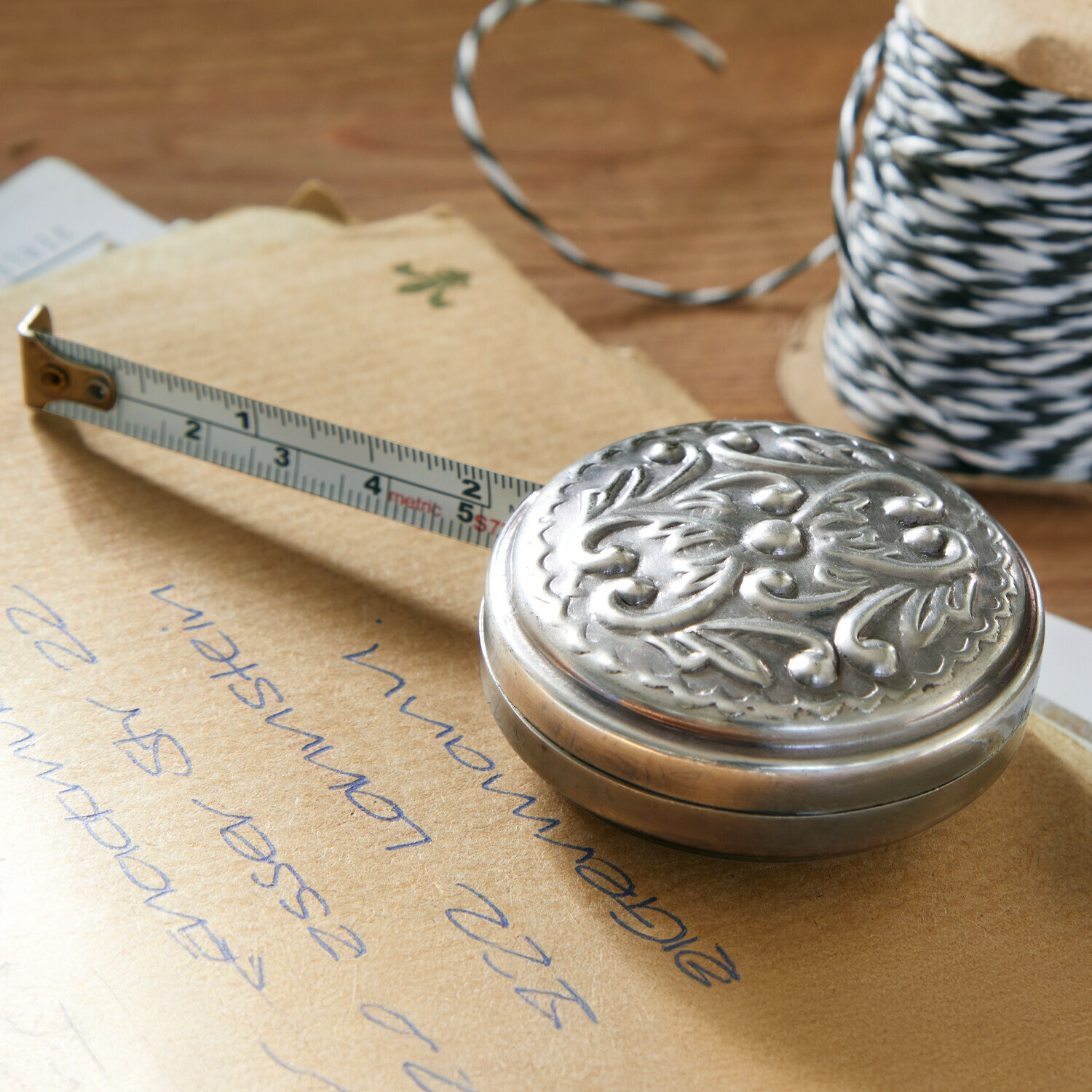 Mètre ruban Dakota Mètre ruban en métal orné de motifs végétaux sur une table en bois avec une bobine de ficelle