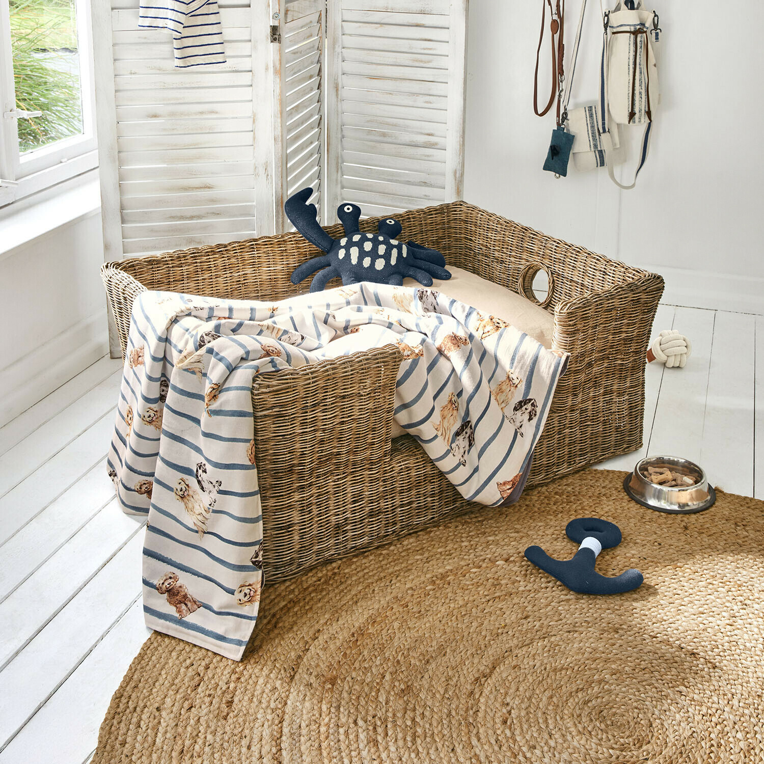 Panier en osier pour chien avec coussin beige et couverture rayée à motif chiens, jouets et gamelle au sol