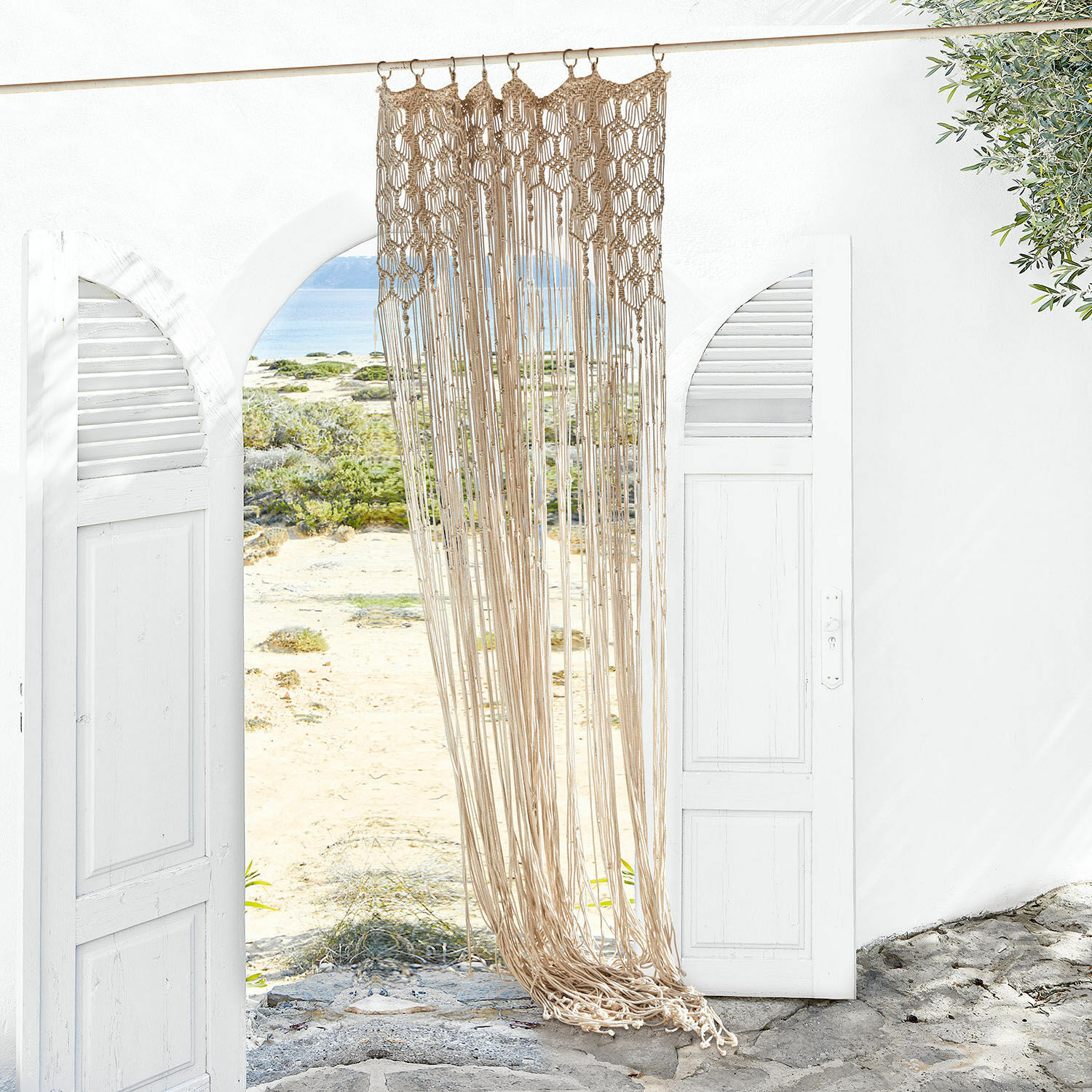 Rideau Manville Macramé beige en coton suspendu dans une entrée blanche ouverte avec vue sur la plage et la mer
