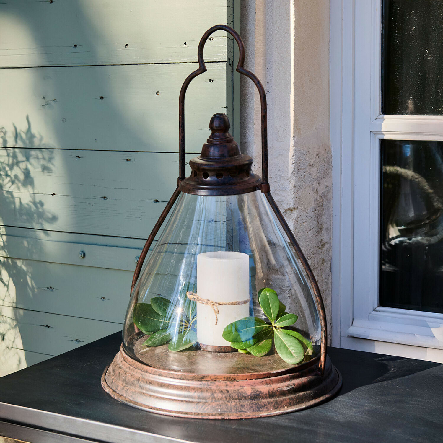 Lanterne en verre avec cadre en métal rouillé, bougie blanche et feuilles vertes sur une table extérieure
