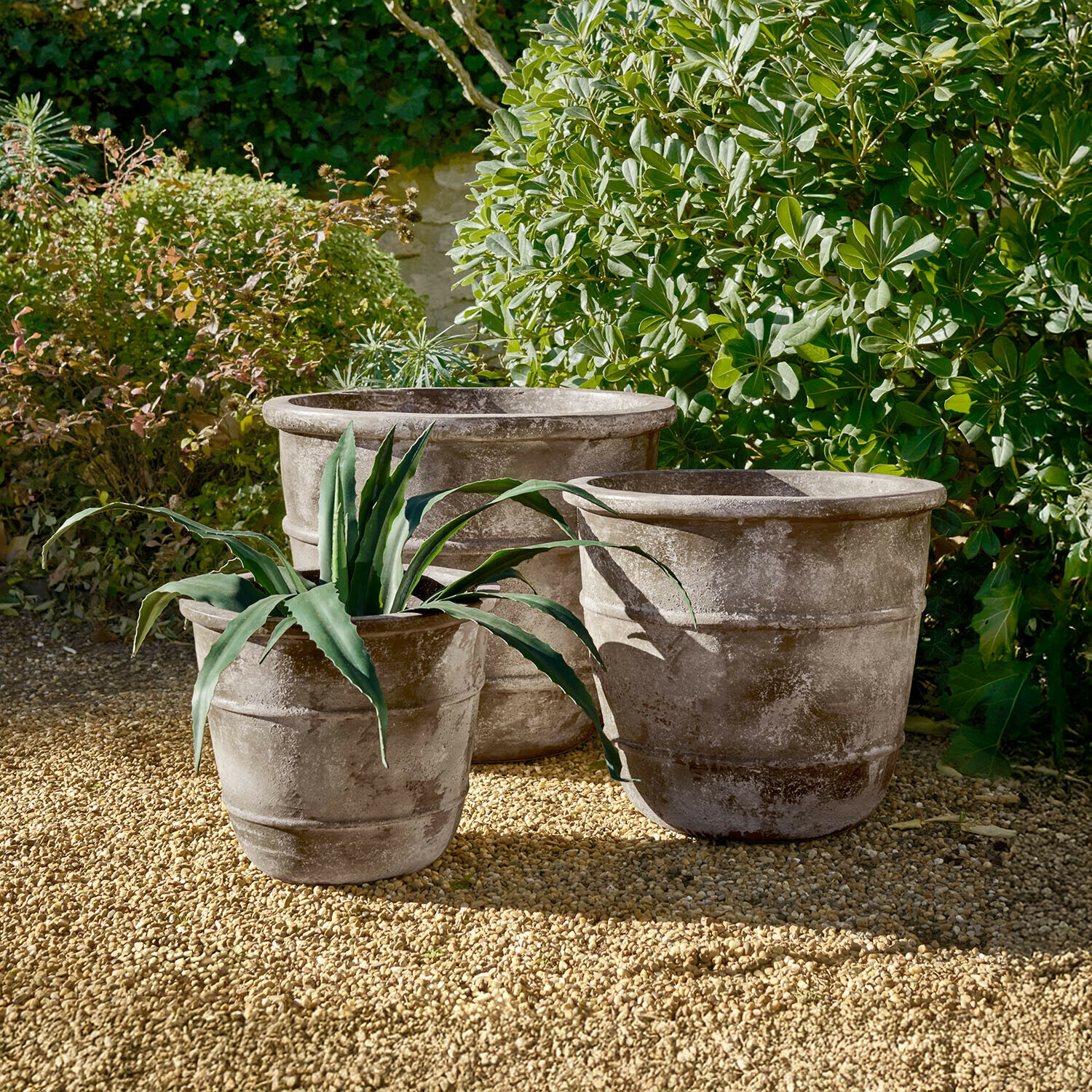 Trois grands pots en pierre grise sur un sol gravillonné dans un jardin avec des plantes vertes