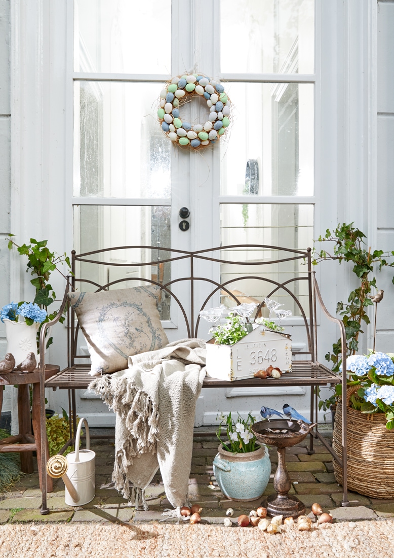 Banc de jardin en métal avec couverture beige et coussin, décorations florales et couronne d'œufs pastel