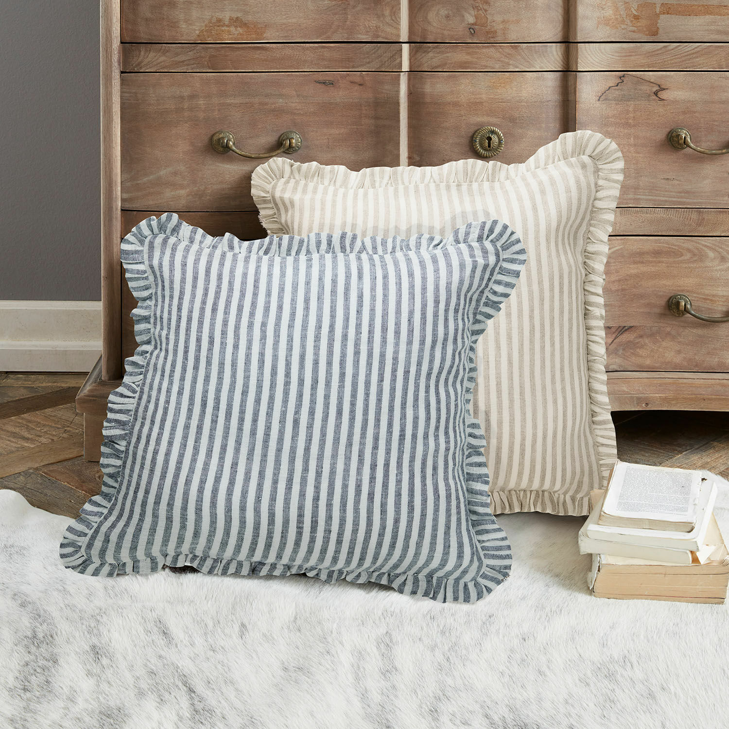 Lot de 2 housses de coussins Brenda Deux coussins décoratifs rayés avec volants beige et bleu posés sur un tapis devant une commode en bois
