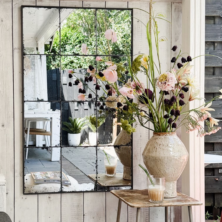 Miroir mural à panneaux reflétant un vase en céramique avec fleurs et un verre de café sur une table en bois rustique