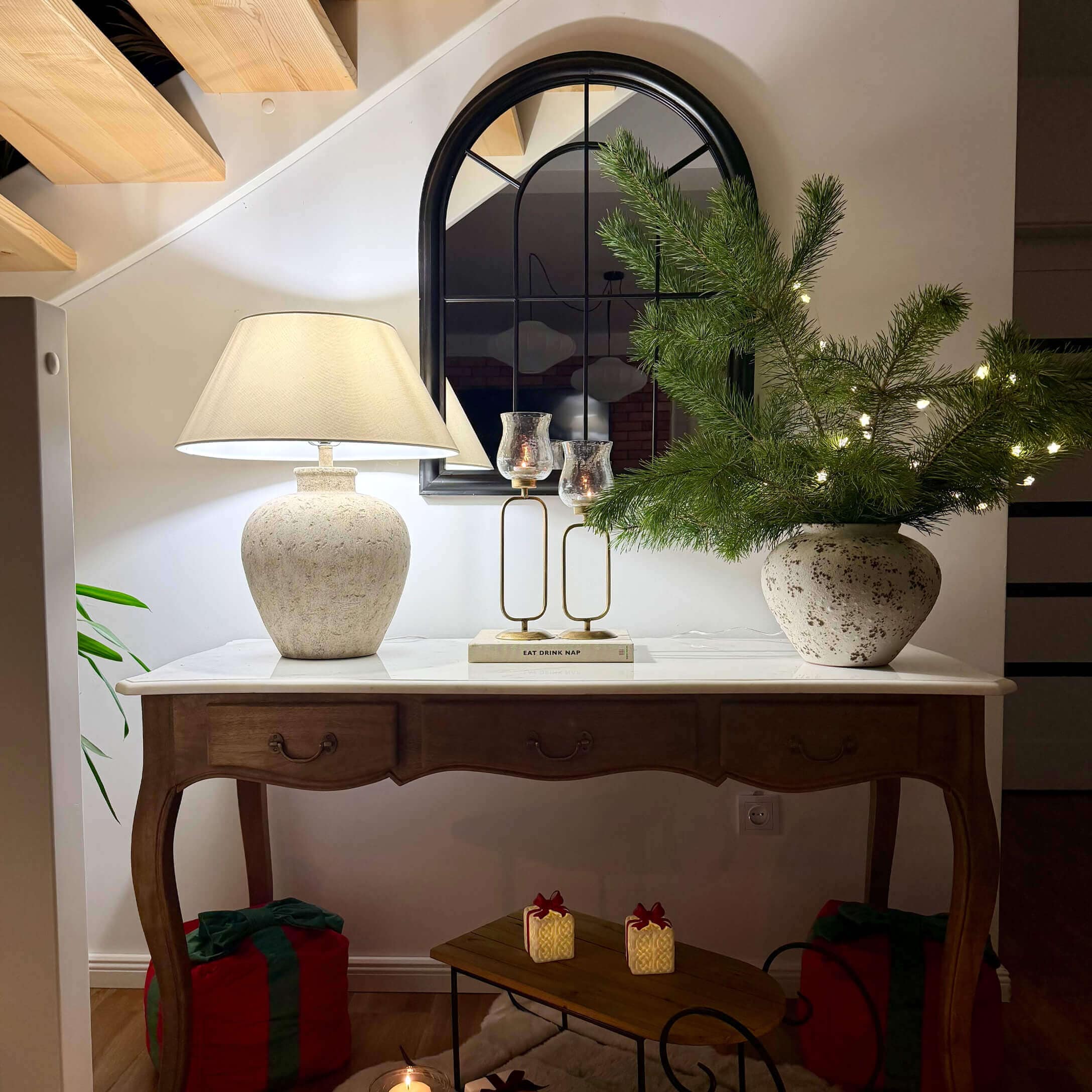 Table en bois avec plateau blanc, lampe en céramique, miroir et décoration de Noël avec branches vertes et lumières