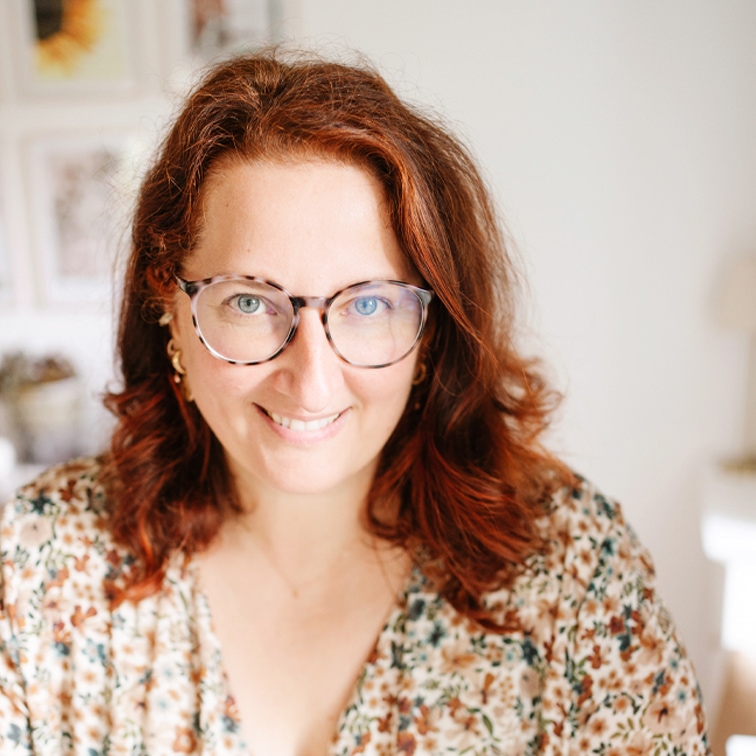 Portrait d'une femme aux cheveux roux et lunettes portant un chemisier fleuri dans un intérieur lumineux