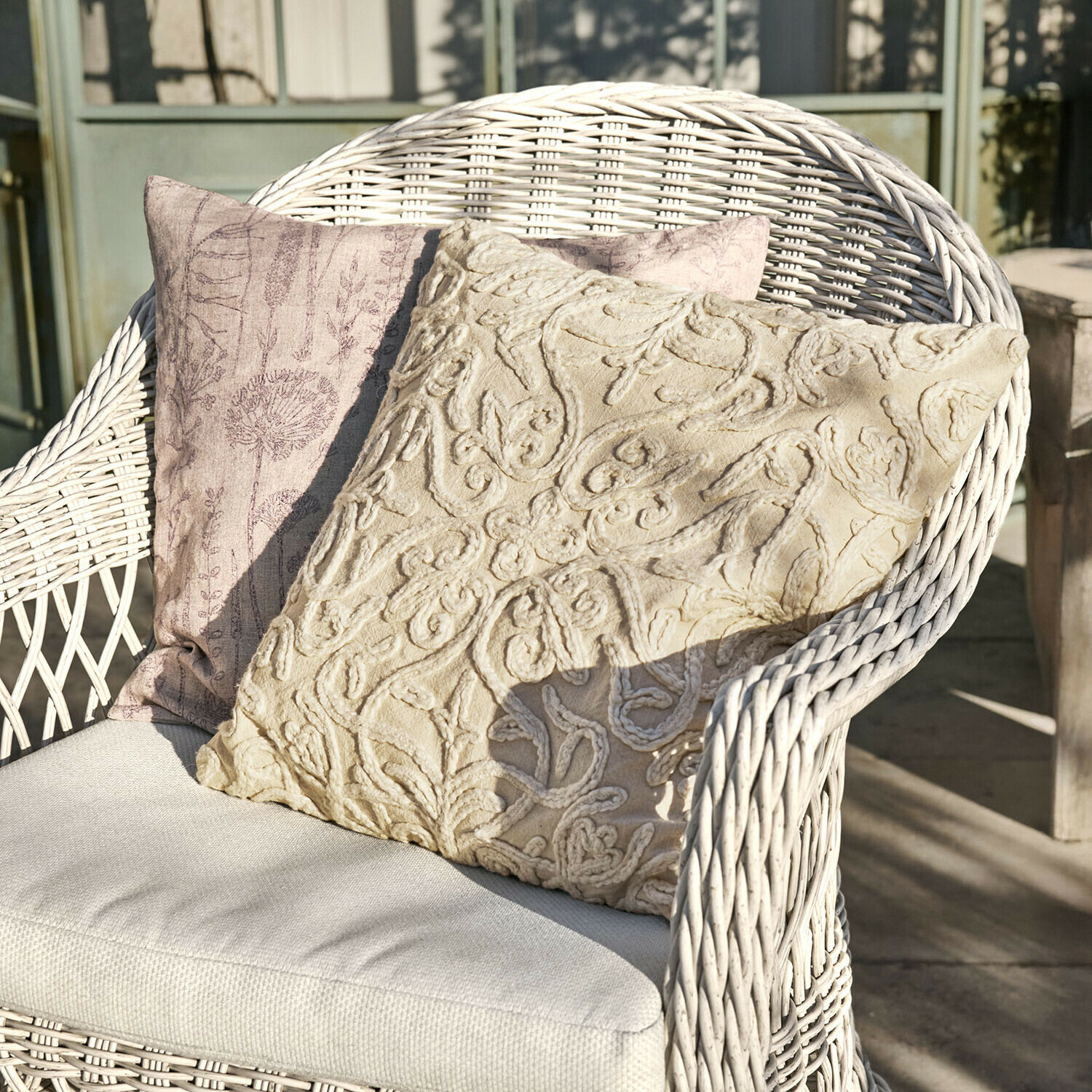 Lot de 2 housses de coussins Floraine Deux coussins décoratifs beige et rose sur un fauteuil en osier blanc sur une terrasse ensoleillée