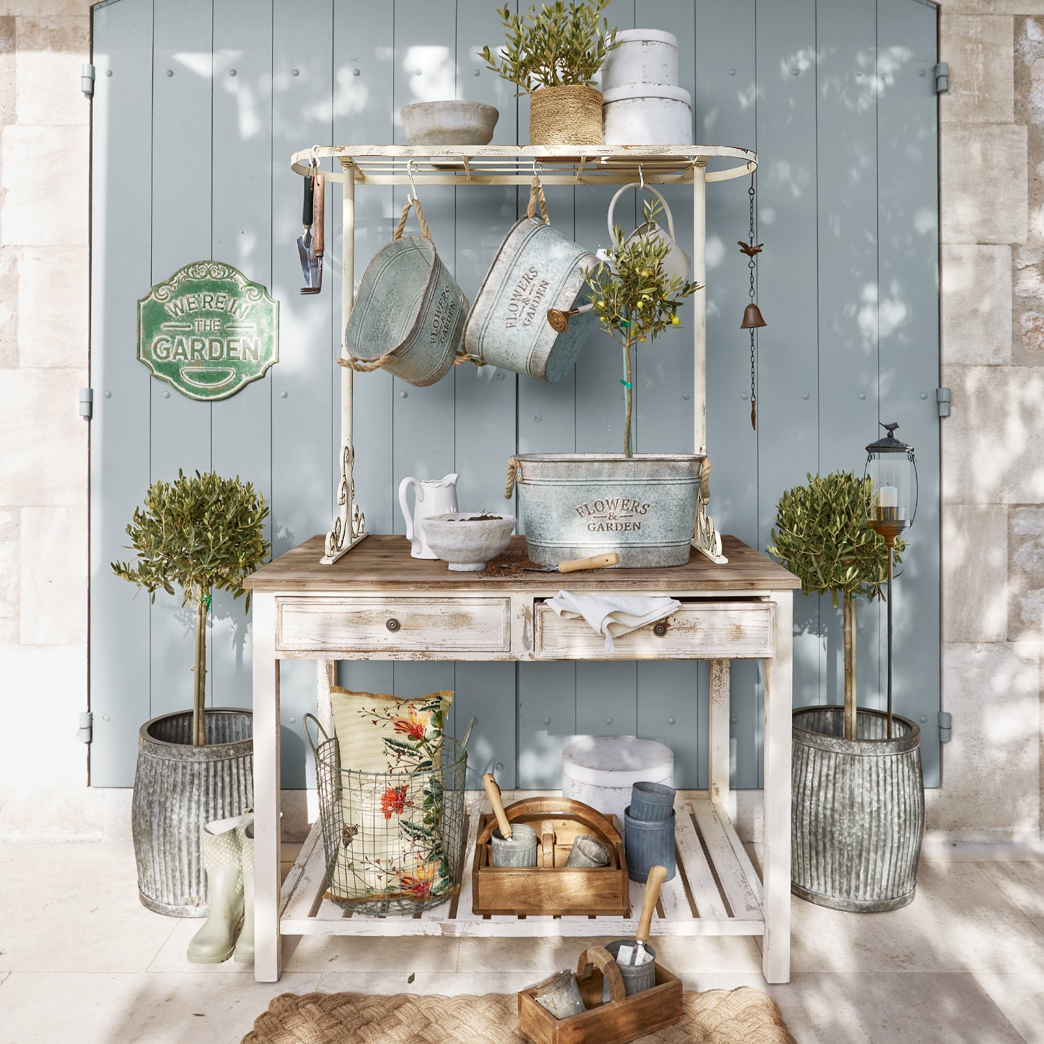 Table de jardin en bois blanc avec pots en métal et outils de jardinage devant une porte bleue