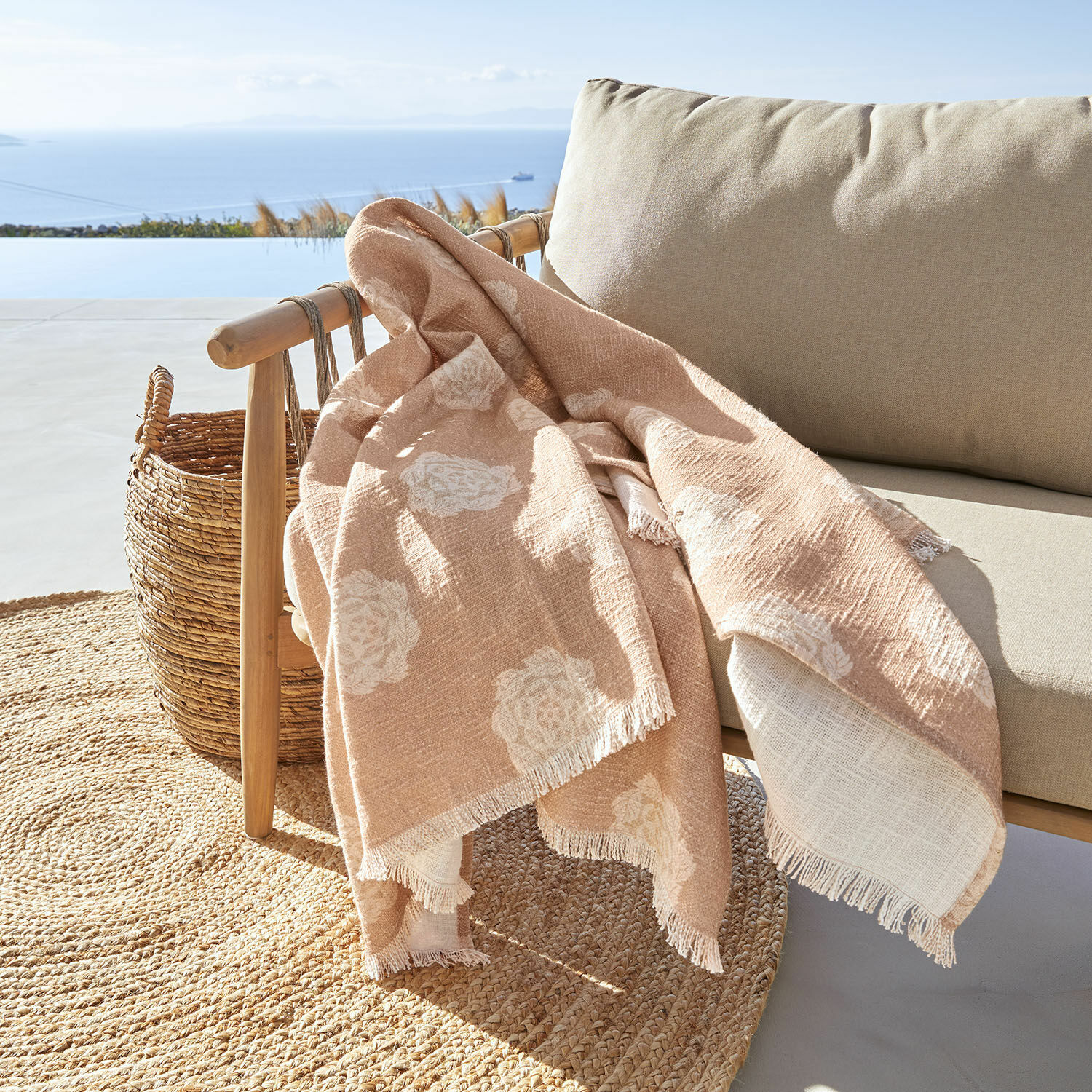 Plaid Belvienne Plaid beige à motif floral blanc en tissu doux sur fauteuil en bois sur terrasse avec vue sur la mer