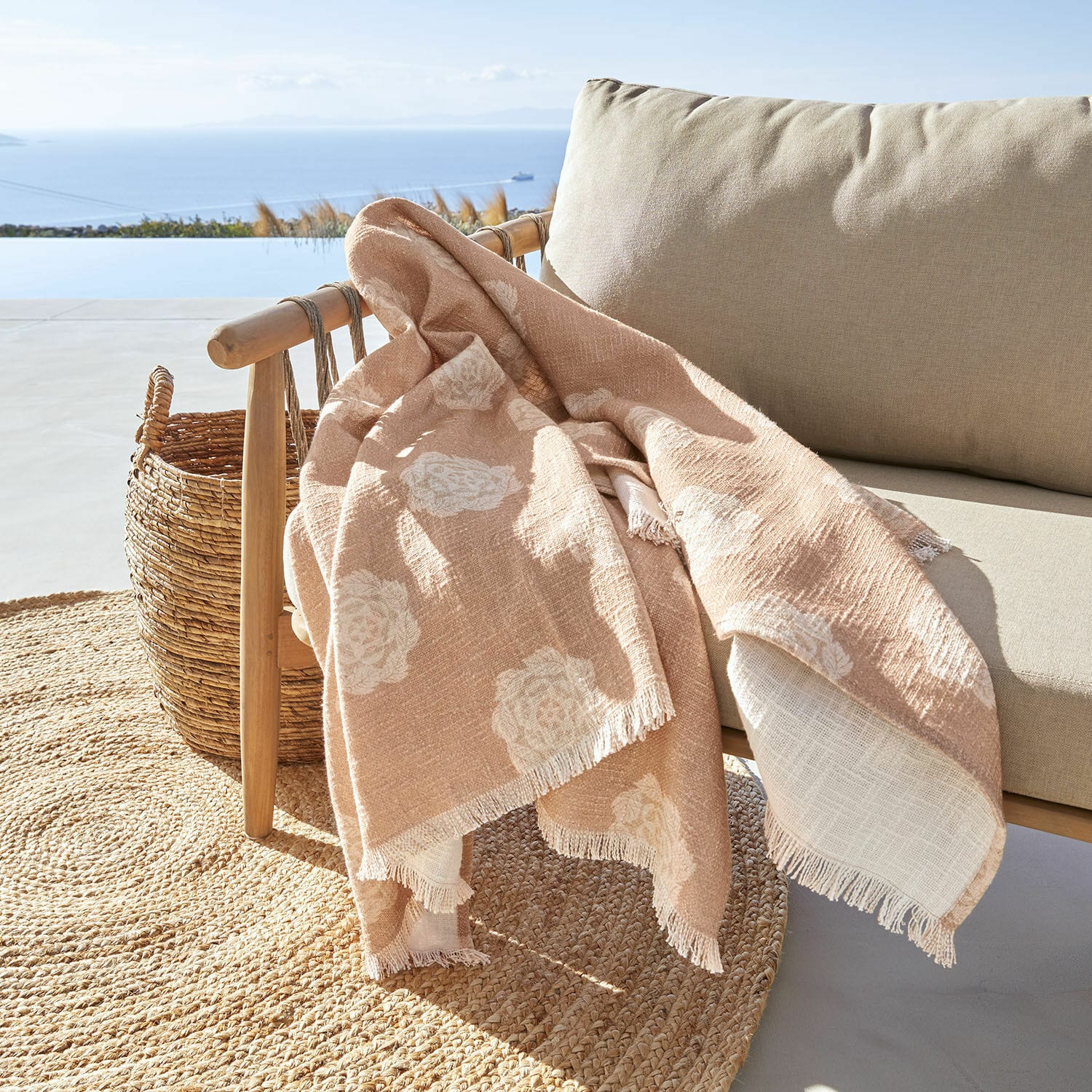 Plaid Belvienne Plaid beige à motif floral blanc en tissu doux sur fauteuil en bois sur terrasse avec vue sur la mer