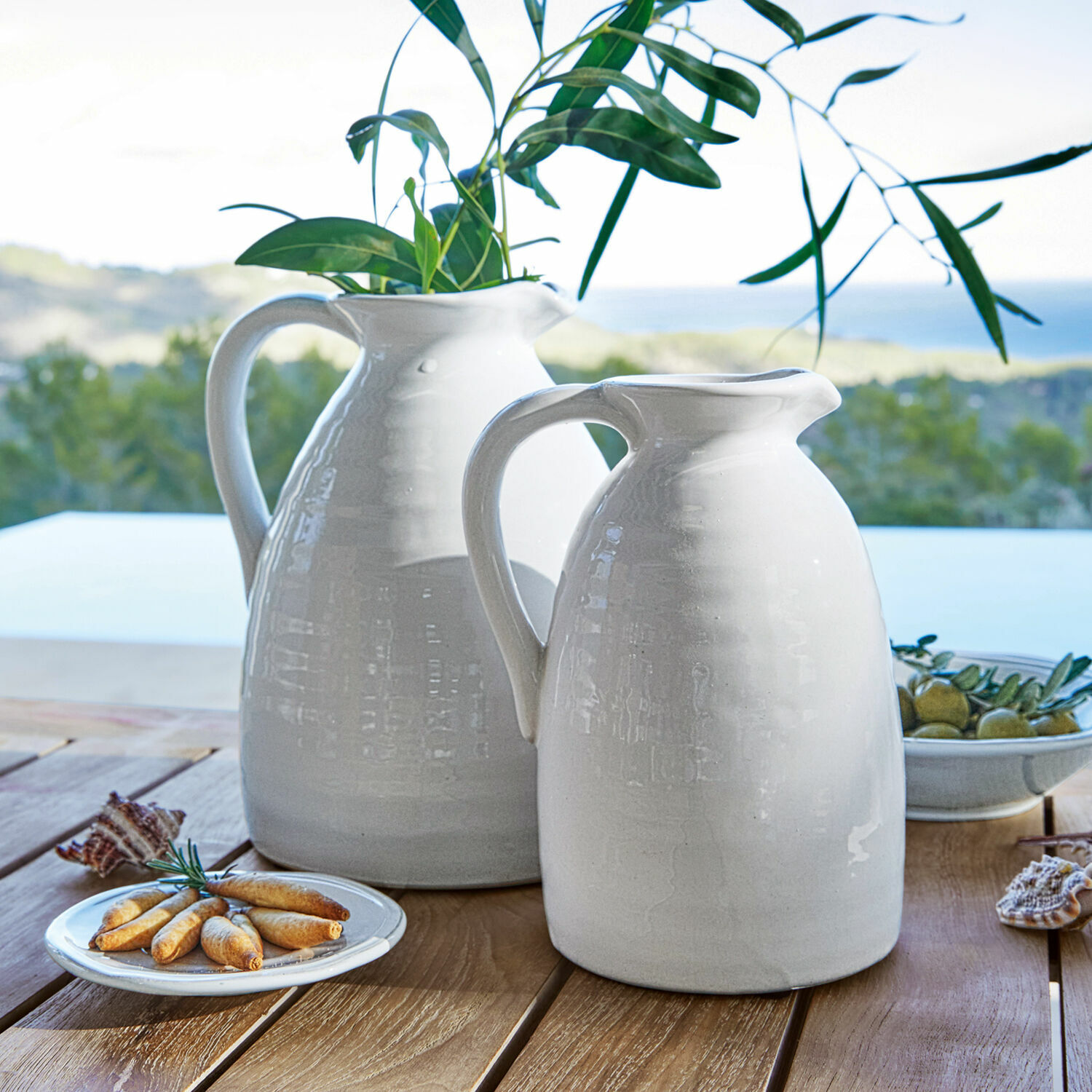 Lot de 2 vases Yarick Carafes blanches en céramique sur table en bois avec feuilles vertes et amuse-bouches, décoration