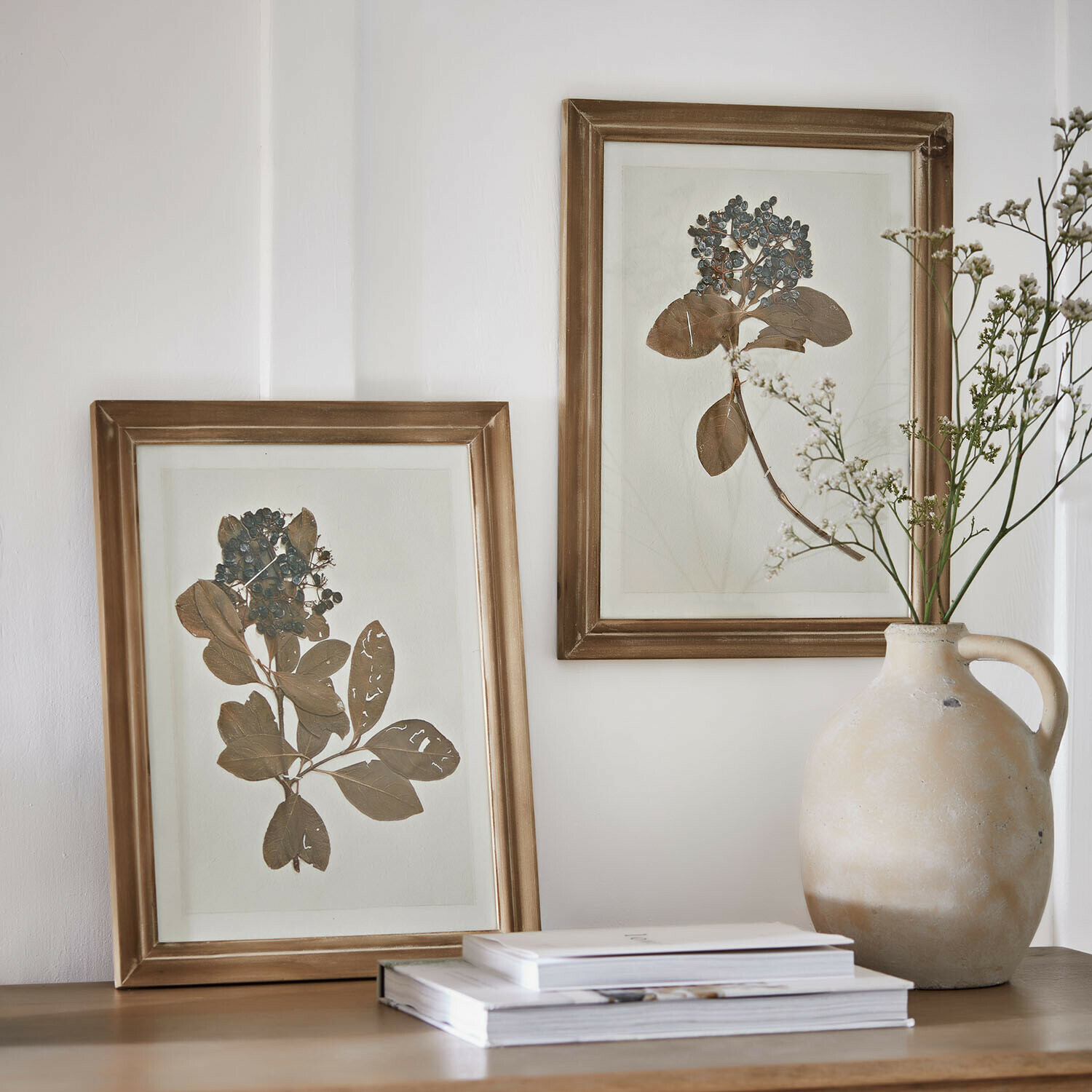 Image set de 2 Tanneau Deux cadres dorés avec feuilles et baies séchées sur une table en bois près d'un vase en céramique et des livres