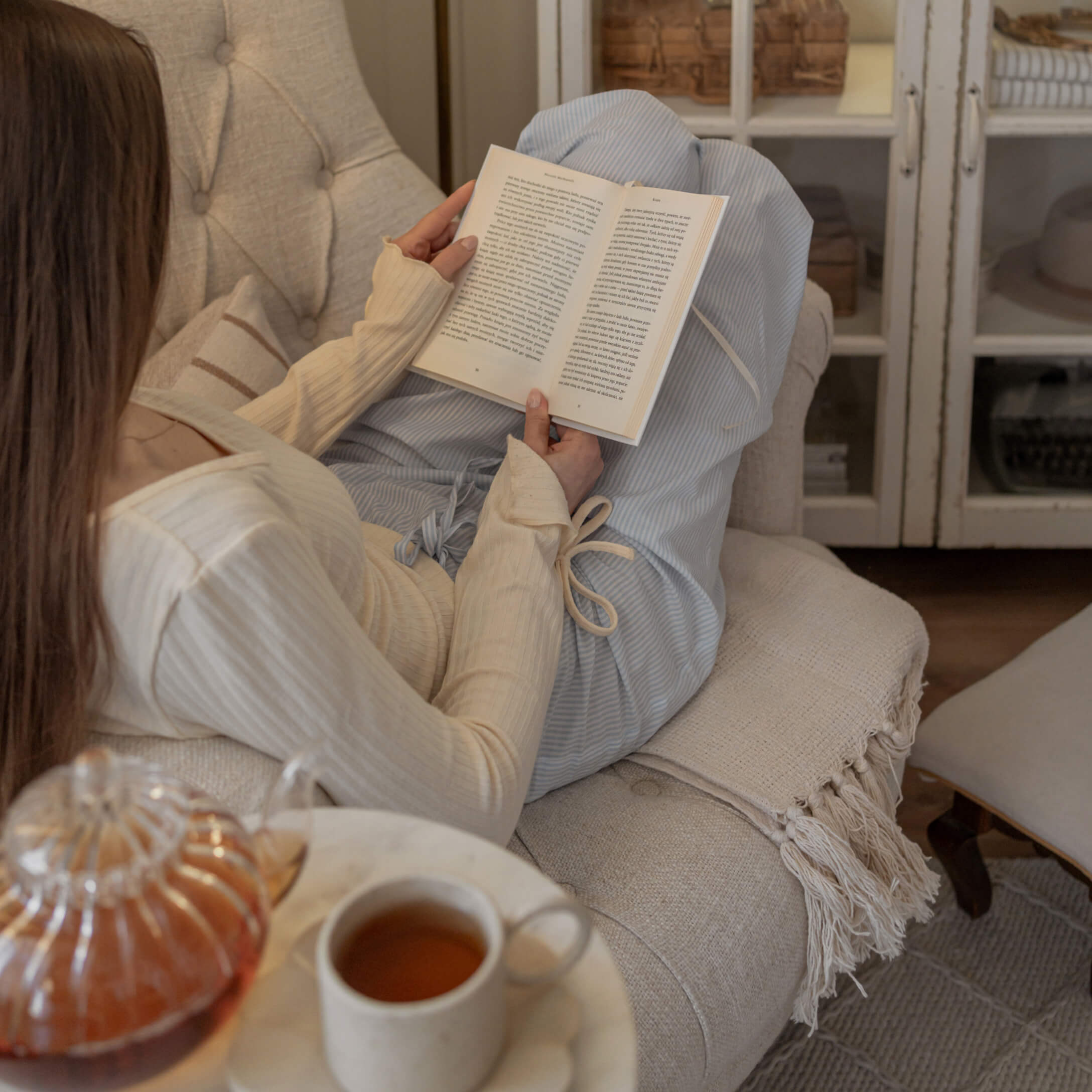 Femme en pull crème et pantalon bleu lisant un livre sur un canapé beige avec une tasse de thé sur la table