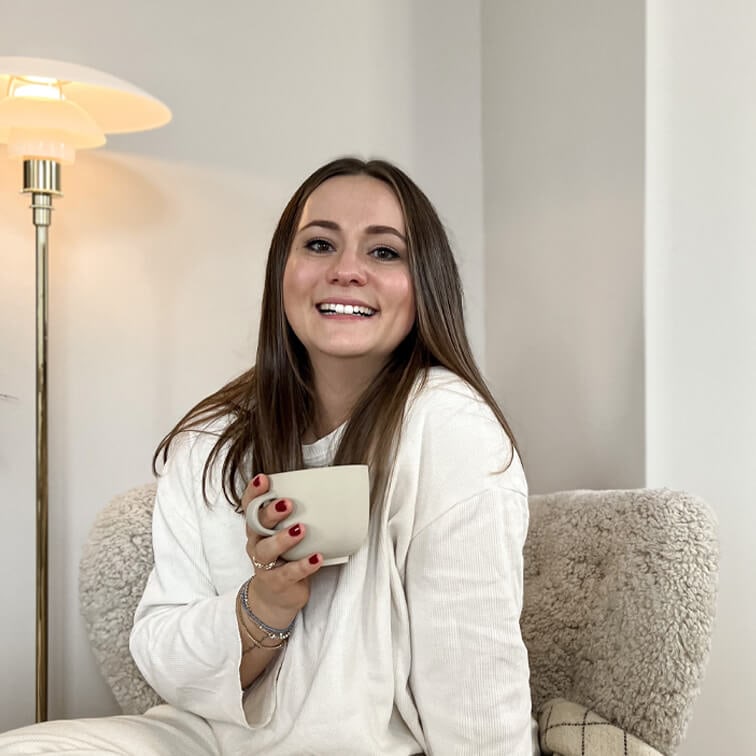 Femme souriante en pull blanc tenant une tasse beige assise sur un fauteuil moelleux dans un intérieur clair