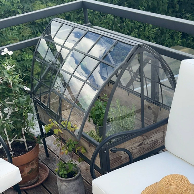 Petite serre de jardin avec cadre en métal et base en bois sur terrasse avec plantes et mobilier extérieur