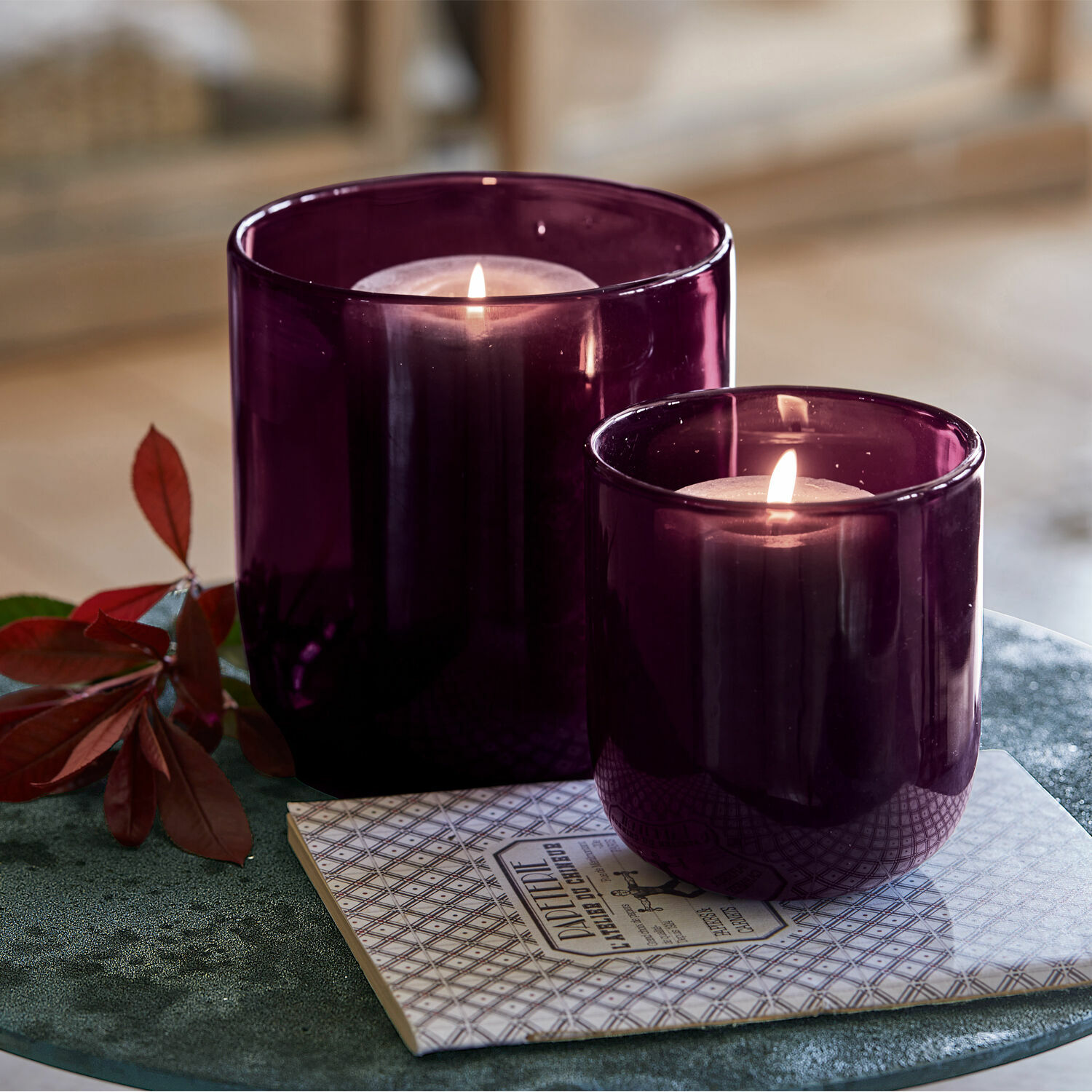 Lot de 2 photophores Theodor Deux photophores en verre violet avec bougies allumées sur un dessous de verre décoratif sur table verte