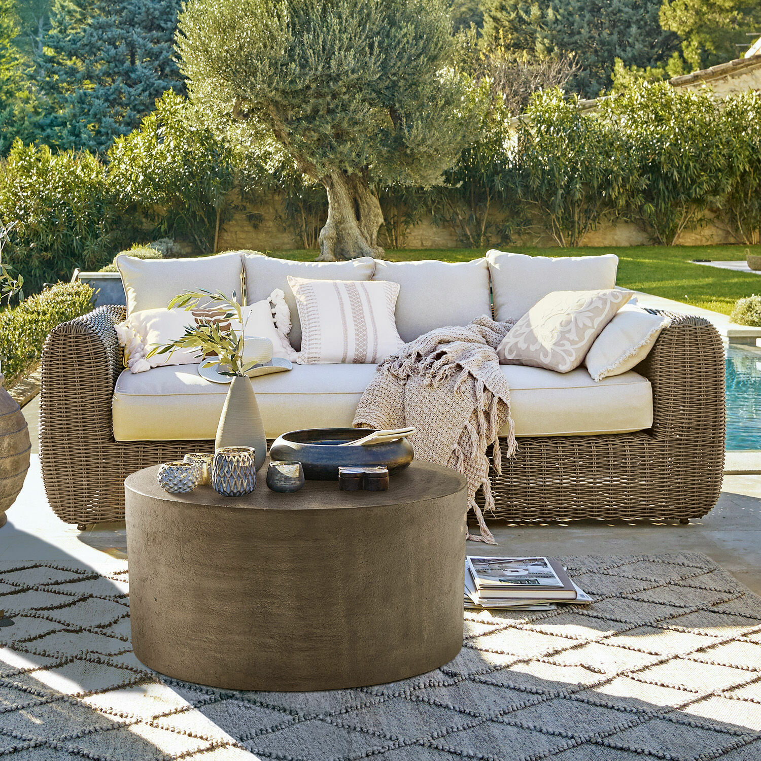 Canapé Bogdanne Canapé d'extérieur en rotin beige avec coussins et table basse ronde en béton dans un jardin