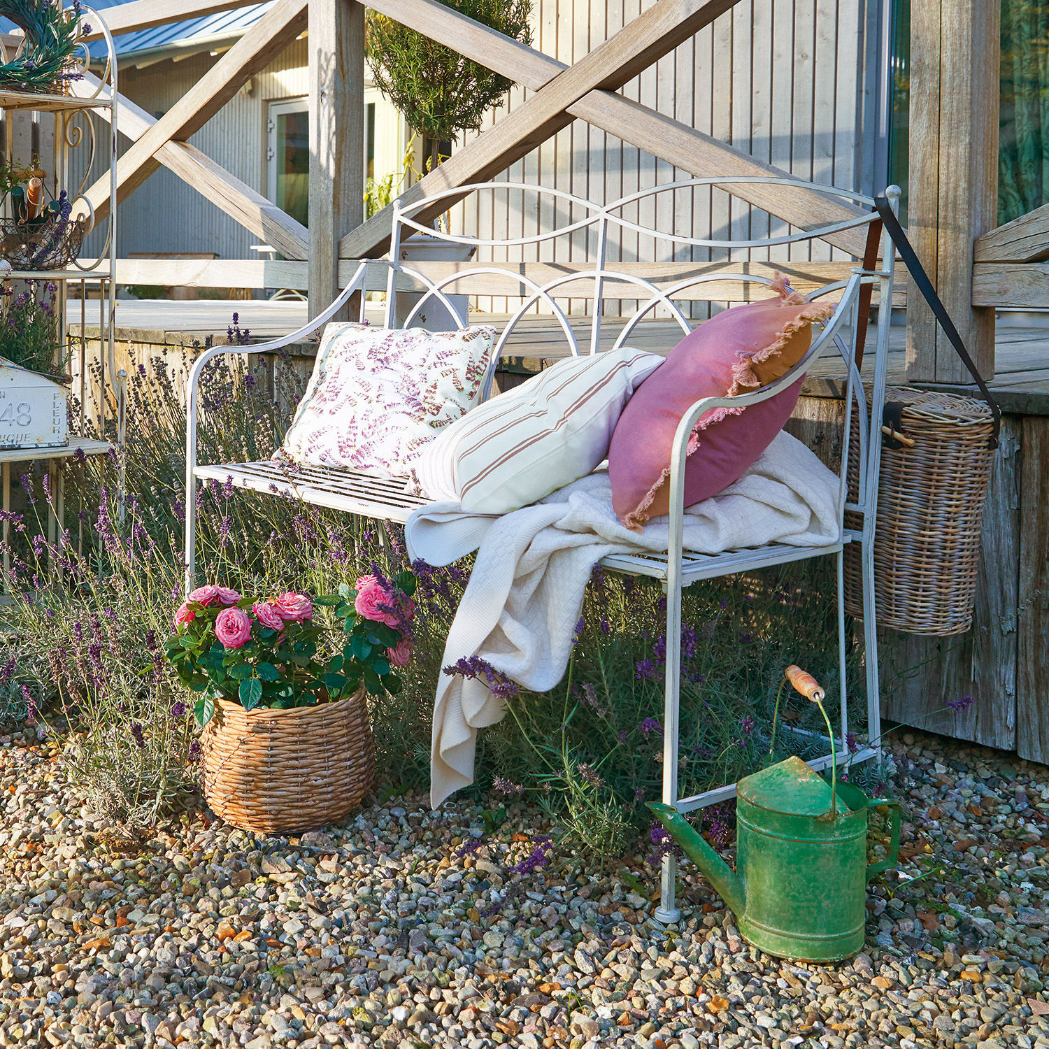 Banc Jablines Banc de jardin en métal blanc avec coussins et couverture sur un chemin de gravier avec fleurs et arrosoir vert