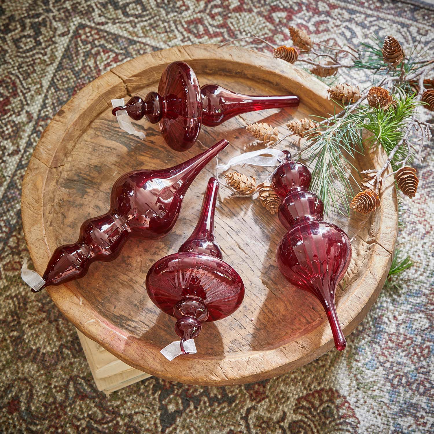 Quatre décorations de Noël en verre rouge sur un plateau en bois avec branches et pommes de pin