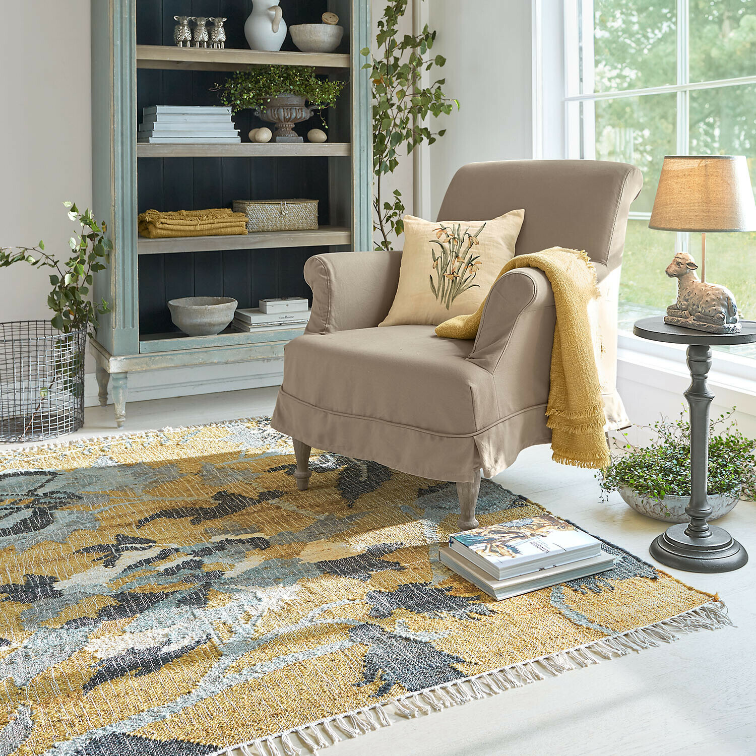 Tapis Ornelless Fauteuil beige avec coussin et plaid jaune sur tapis coloré dans un salon lumineux avec étagère