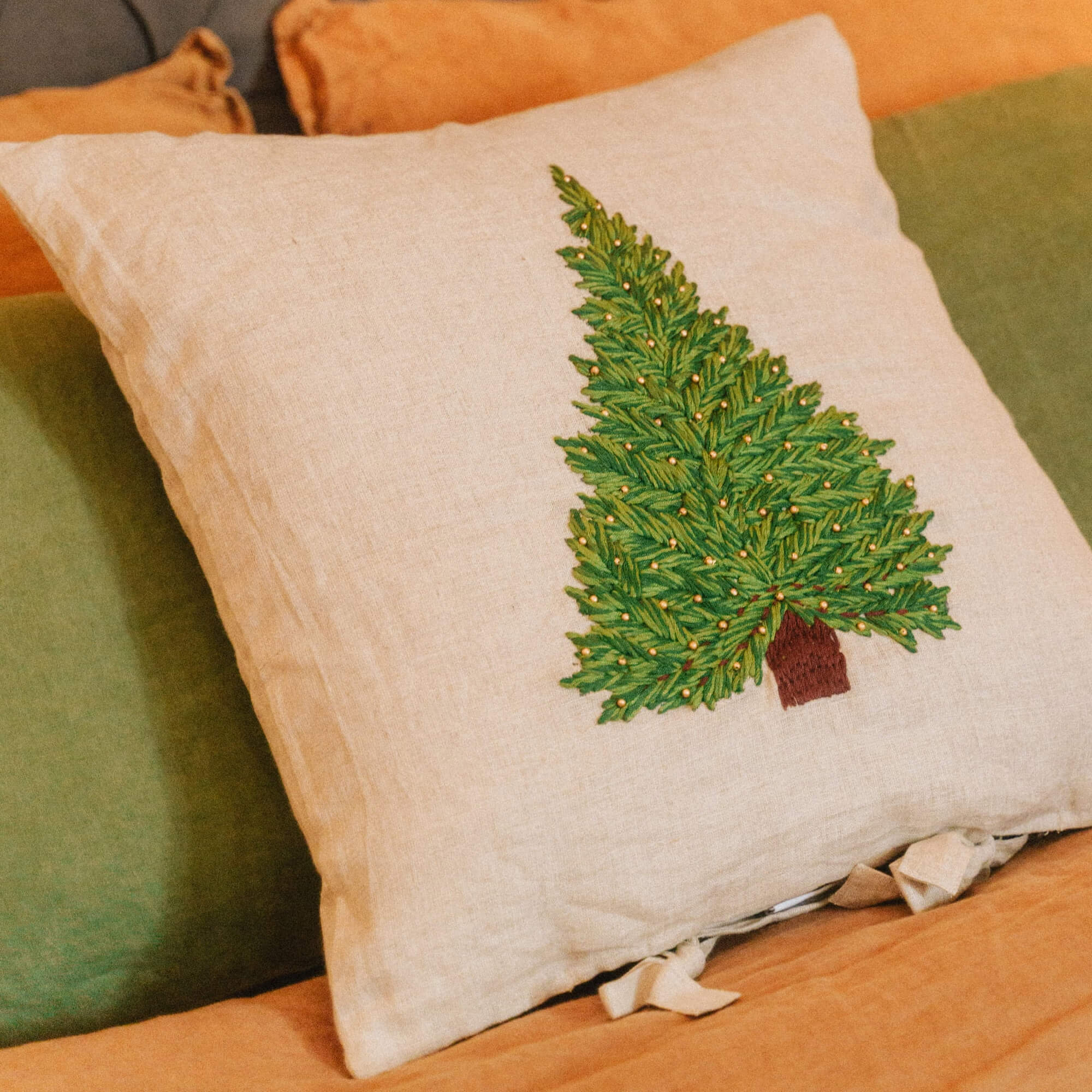 Coussin en lin beige avec broderie d'arbre de Noël vert et accents dorés sur lit orange et vert