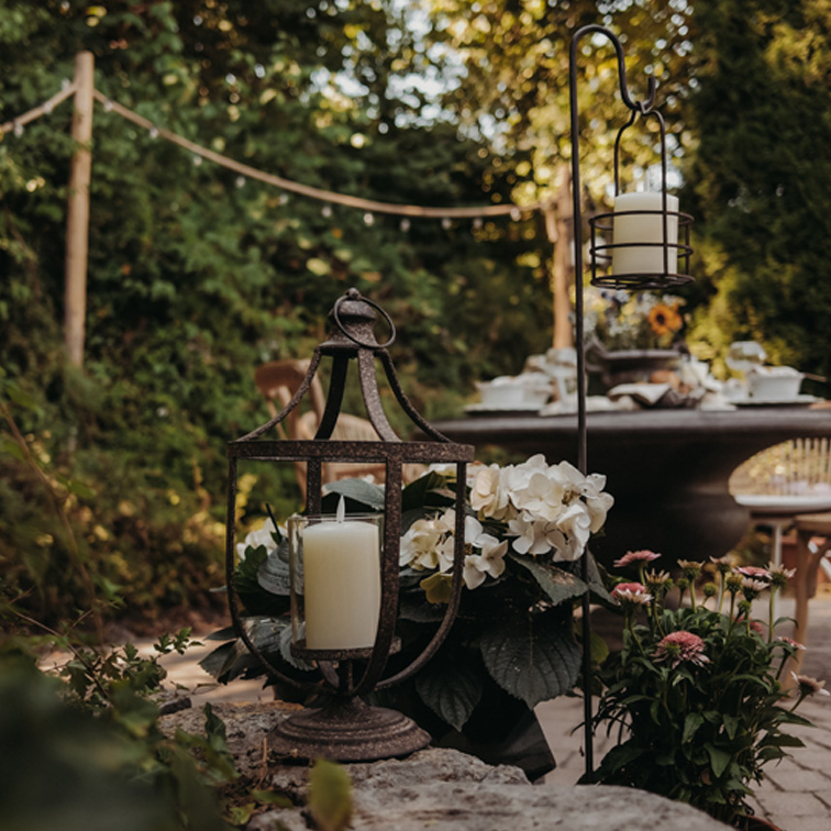 Lanterne en métal avec bougie blanche sur un muret en pierre dans un jardin fleuri avec table en arrière-plan