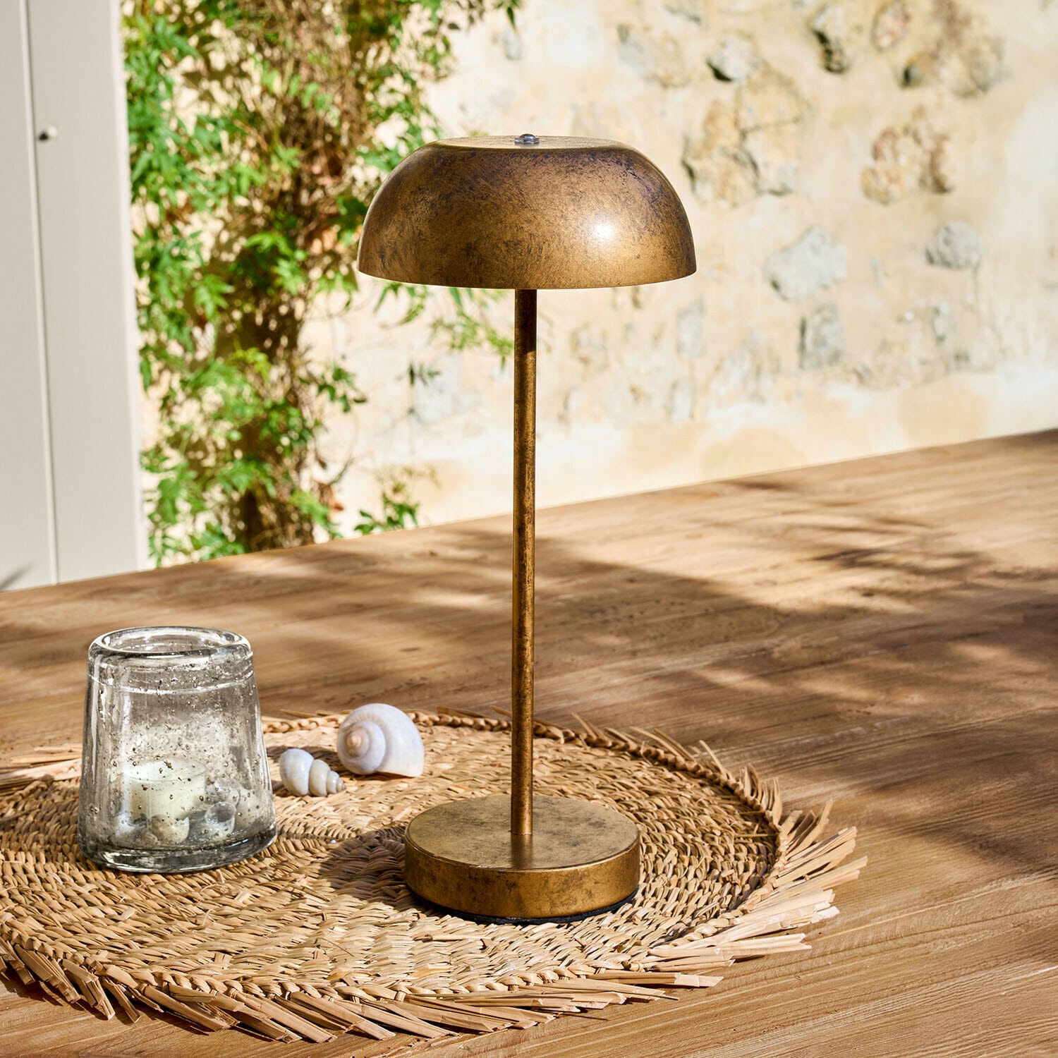 Lampe à poser rechargeable Lumirah Lampe de table en métal couleur or ancien posée sur un dessous de verre tressé sur une table en bois