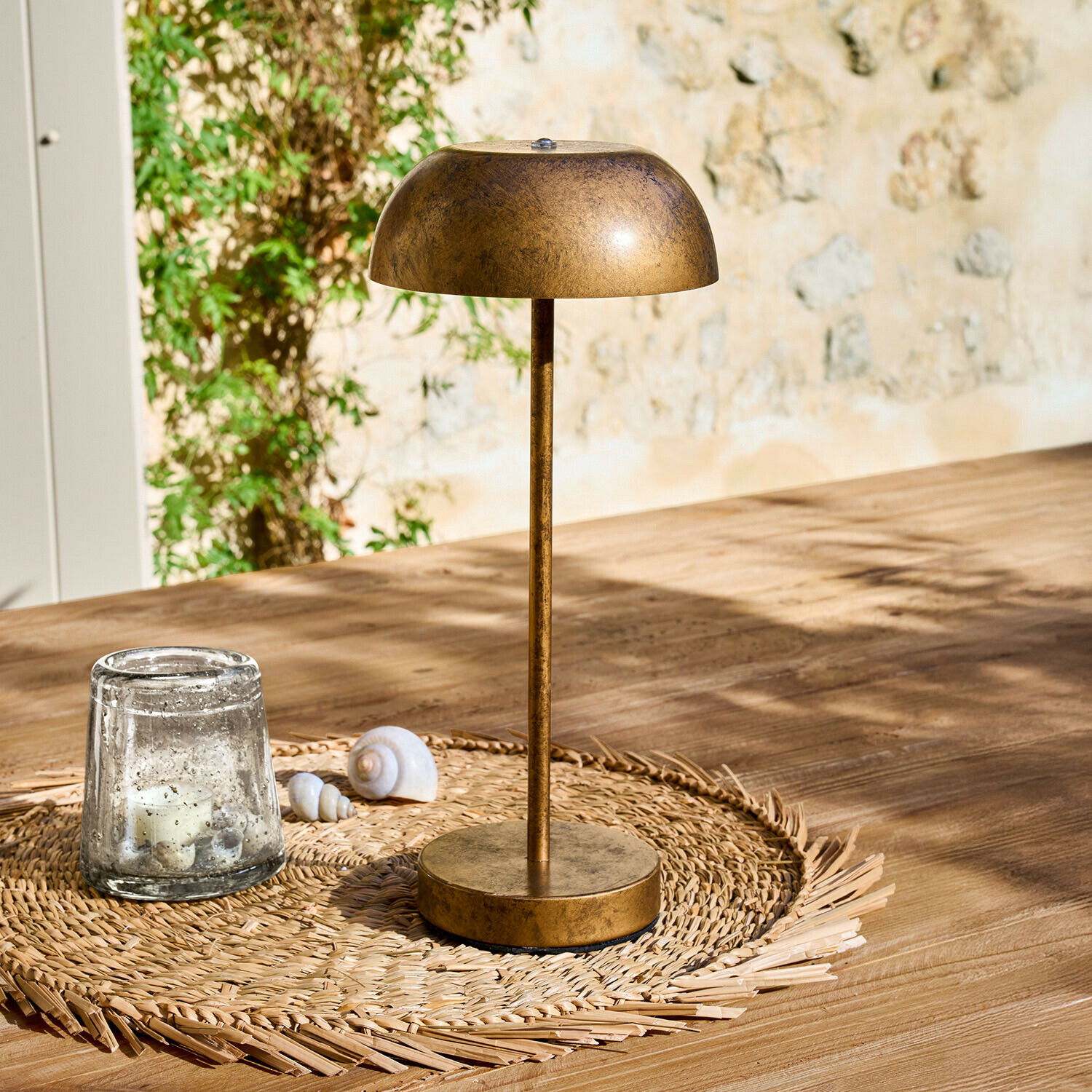 Lampe à poser rechargeable Lumirah Lampe de table en métal couleur or ancien posée sur un dessous de verre tressé sur une table en bois