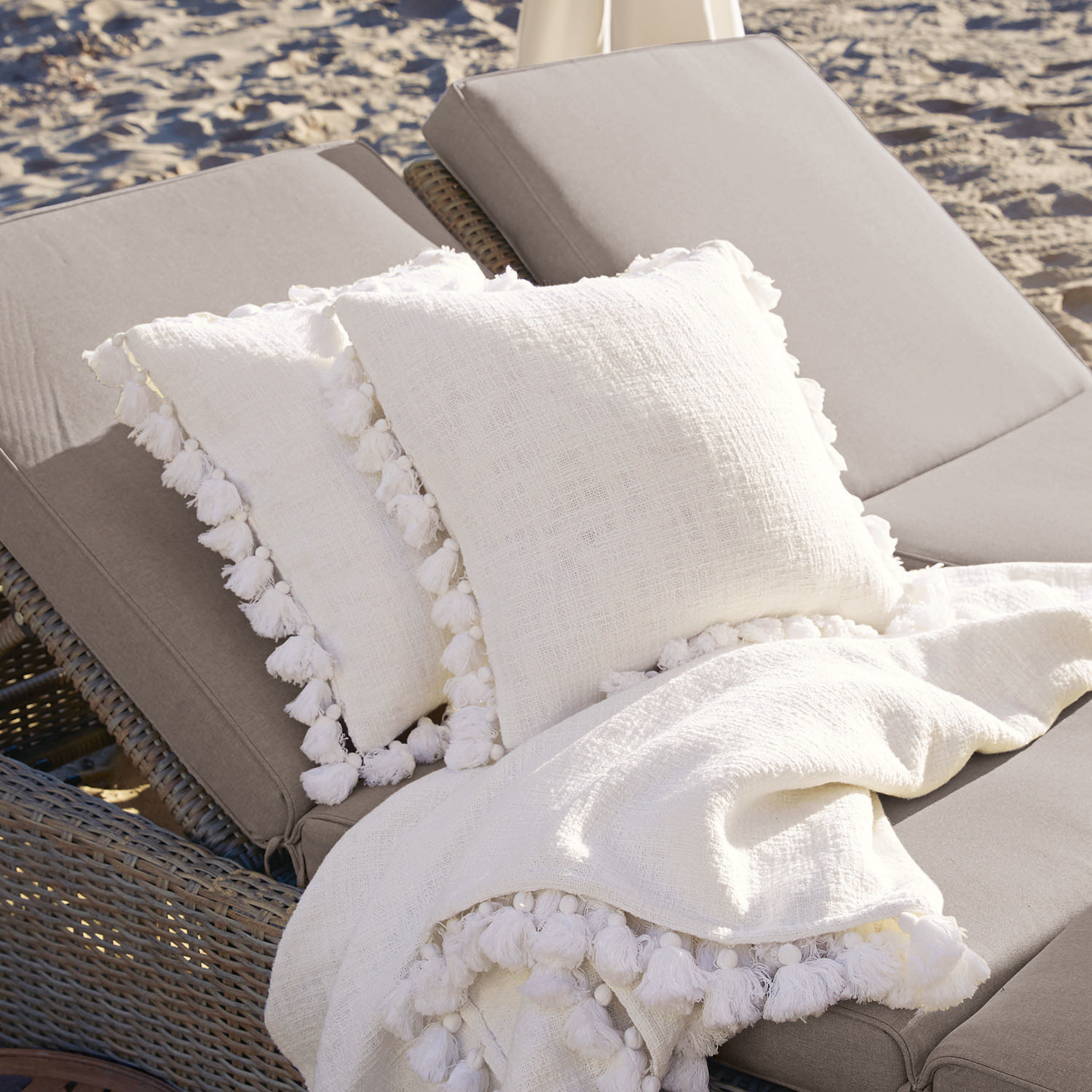 Lot de 2 housses de coussins Peniel Coussins et plaid blancs en lin avec pompons sur chaise longue grise au bord de la plage
