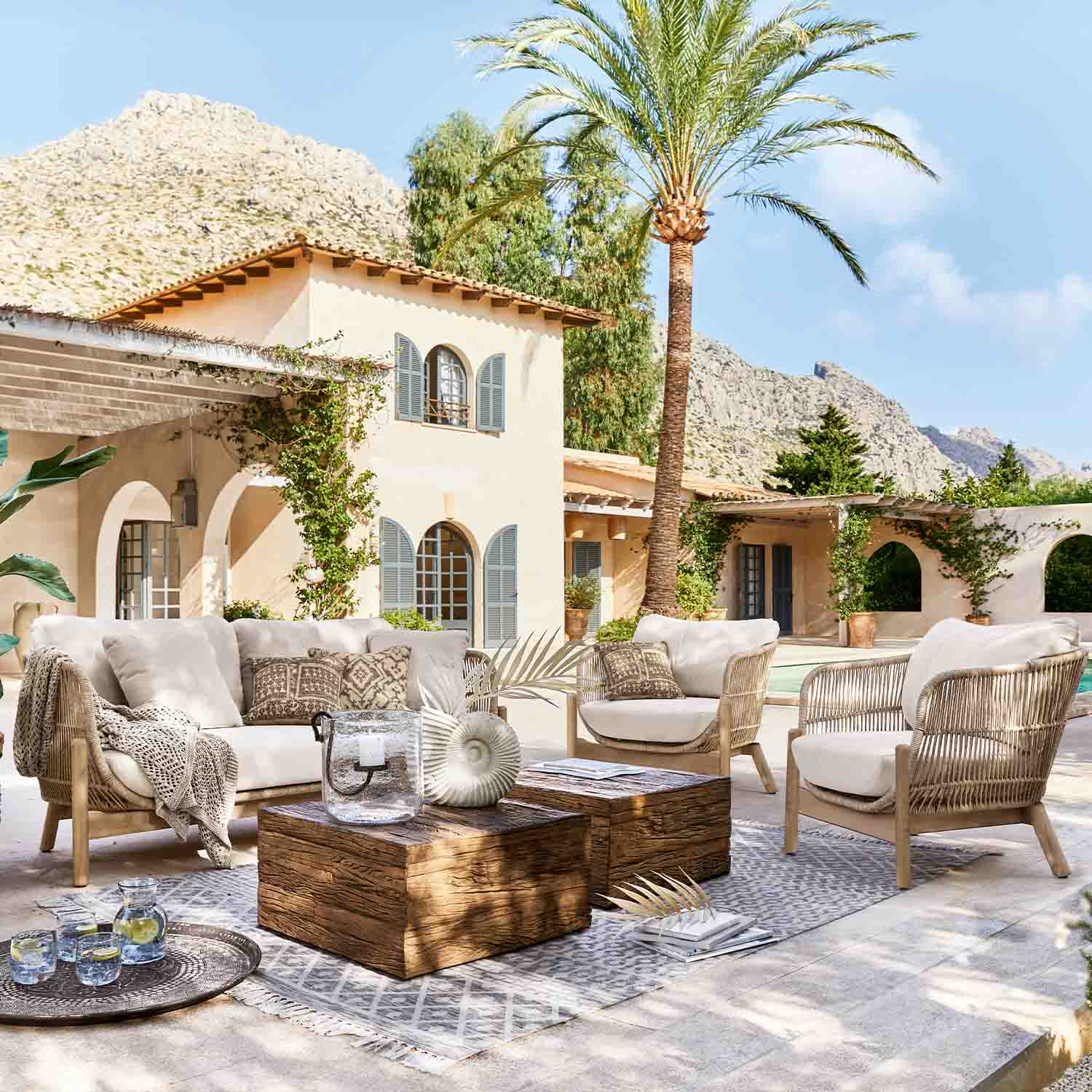 Ensemble de mobilier d'extérieur en rotin clair avec coussins beige sur terrasse avec palmier et montagnes en arrière-plan