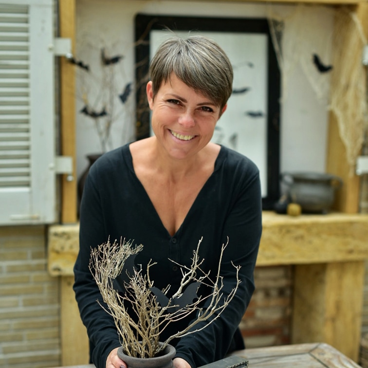 Femme en haut noir tenant un pot avec des branches sèches et décorations chauves-souris dans un intérieur rustique