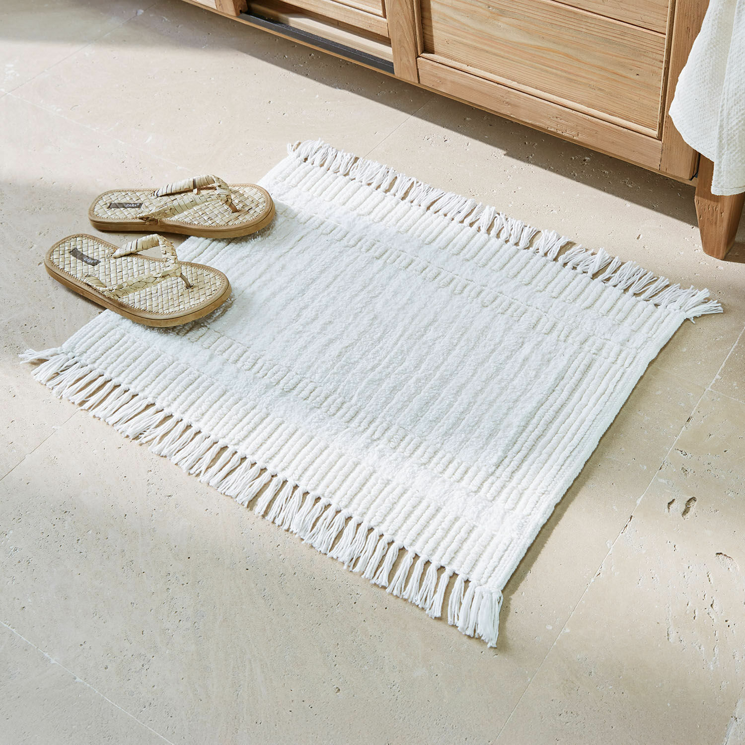 Tapis de bain Nexilo Tapis de bain blanc en coton avec franges sur sol beige et tongs en osier