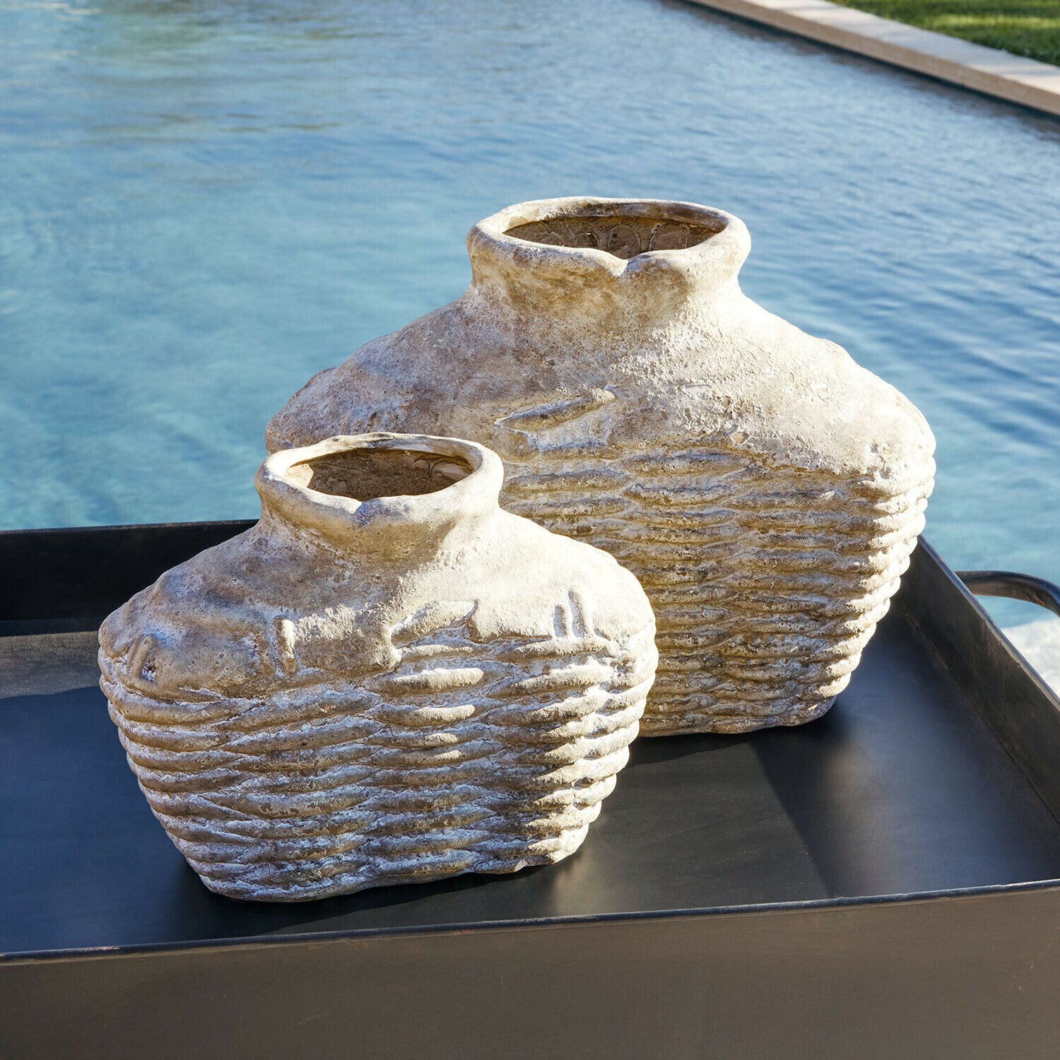 Lot de 2 vases Orlaithea Deux vases décoratifs en pierre beige posés sur un plateau noir près d'une piscine