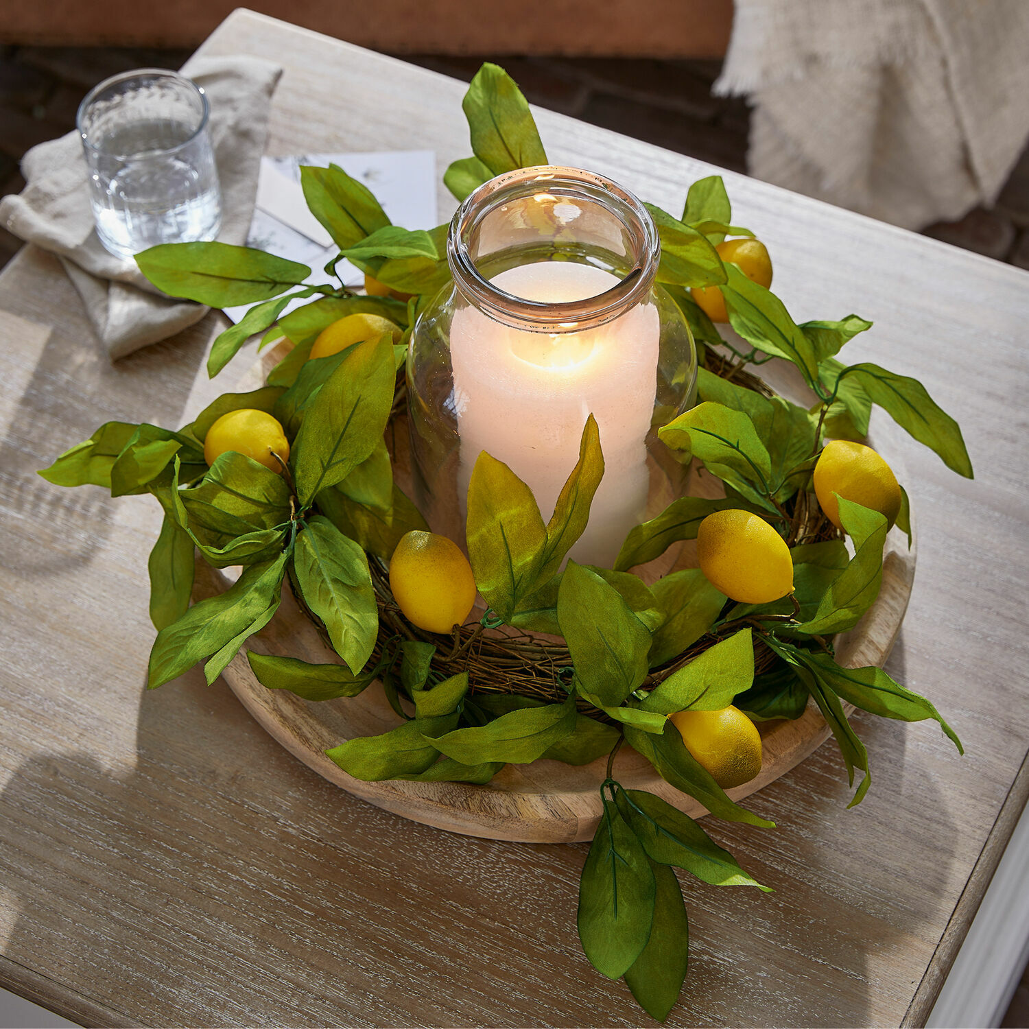 Couronne Lantagenet Bougeoir en verre avec bougie blanche entouré de feuilles vertes et fruits jaunes sur plateau en bois