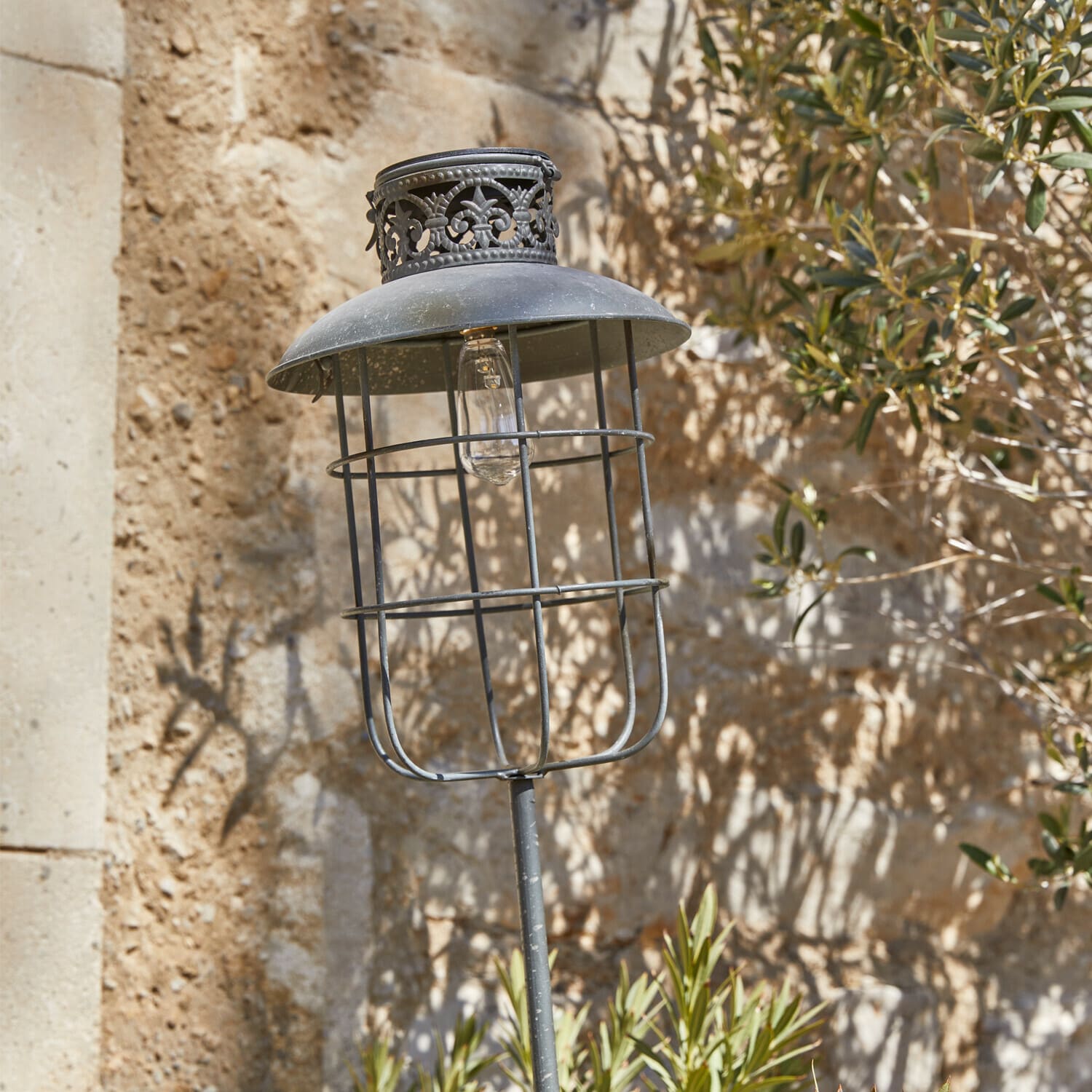 Lampe de jardin en métal noir avec abat-jour décoratif et ampoule visible sur fond de mur en pierre