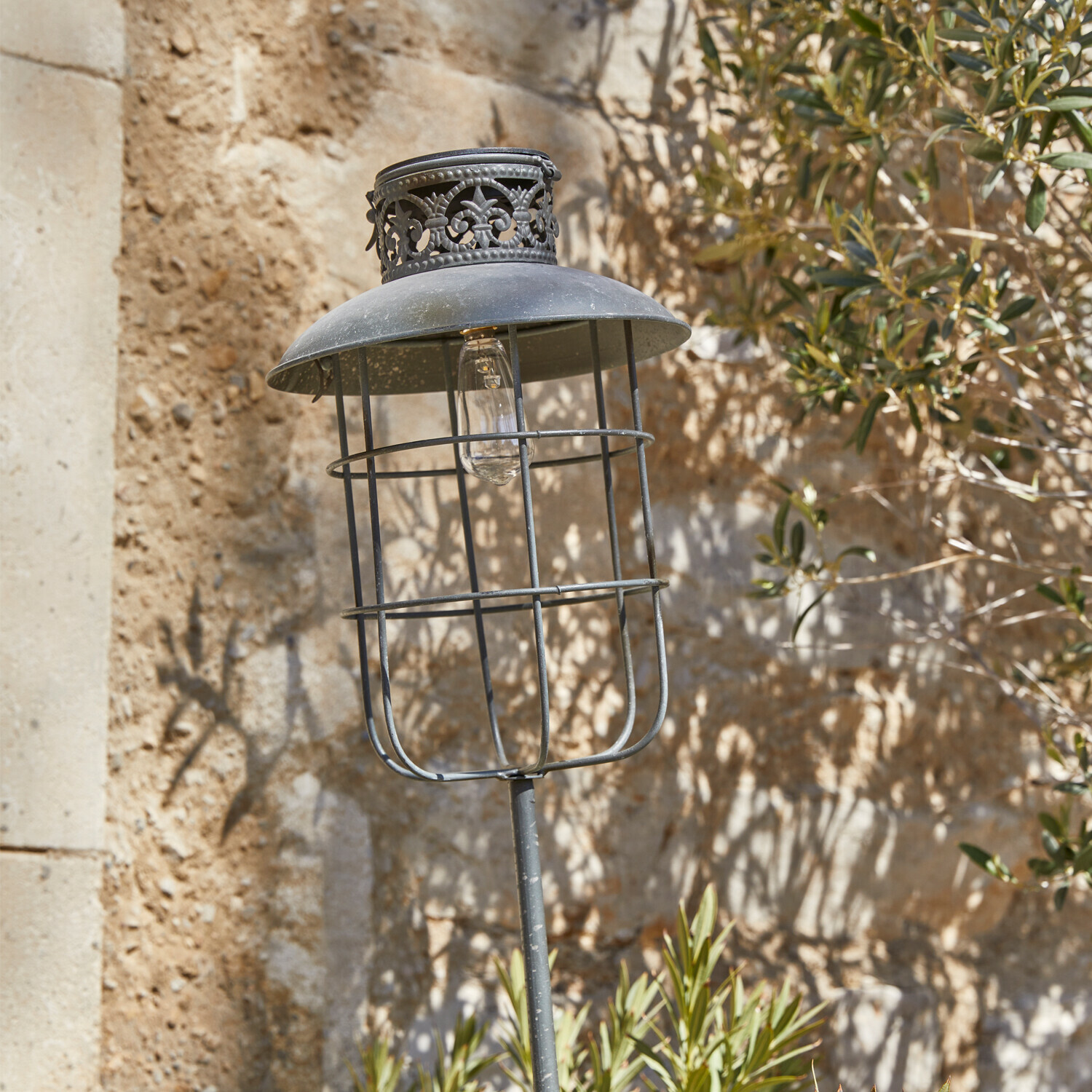 Lanterne solaire Nistos Lampe de jardin en métal noir avec abat-jour décoratif et ampoule visible sur fond de mur en pierre