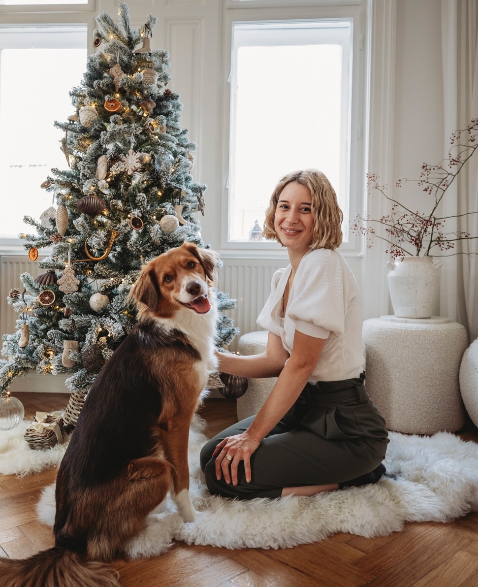 Femme en haut blanc et pantalon vert avec chien près d'un sapin de Noël décoré dans une pièce lumineuse