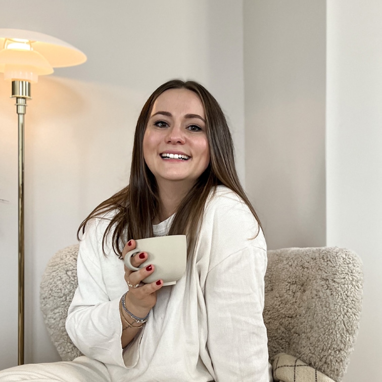Femme souriante en pull blanc tenant une tasse beige assise dans un fauteuil moelleux dans un intérieur clair